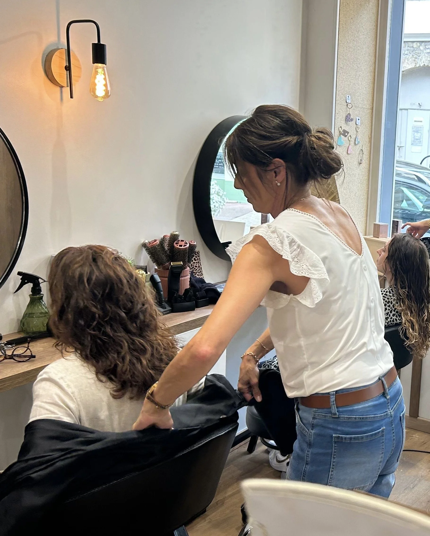 Une femme se tient devant un miroir dans un salon de beauté ou un salon de coiffure, entourée de personnes. La femme porte une chemise blanche avec des détails en dentelle et des jeans. La pièce est décorée de manière moderne avec un éclairage chaud et quelques objets de maquillage ou de coiffure sur la table devant elle.