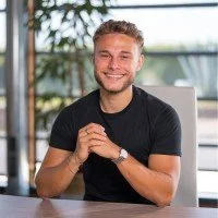 Jeune homme souriant assis à un bureau avec un ordinateur portable, dans un environnement lumineux et moderne.