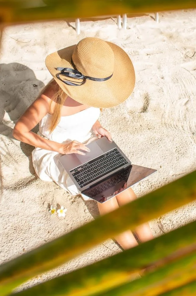 Femme assise sur la plage avec un grand chapeau en paille, utilisant un ordinateur portable.