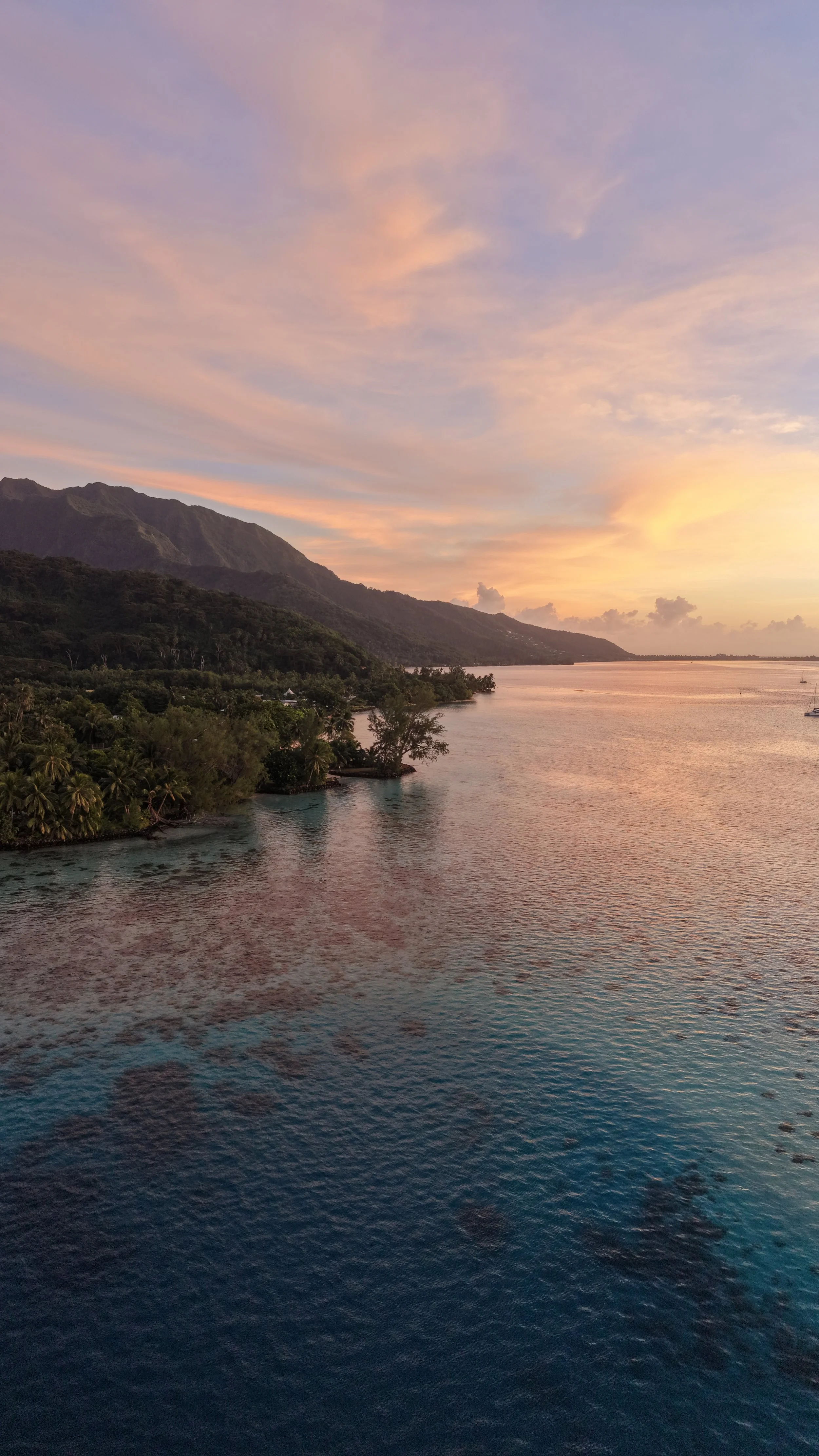 Coucher de soleil sur une mer calme avec des montagnes en arrière-plan, ciel aux teintes roses, oranges et violettes, quelques nuages et un voilier au loin.