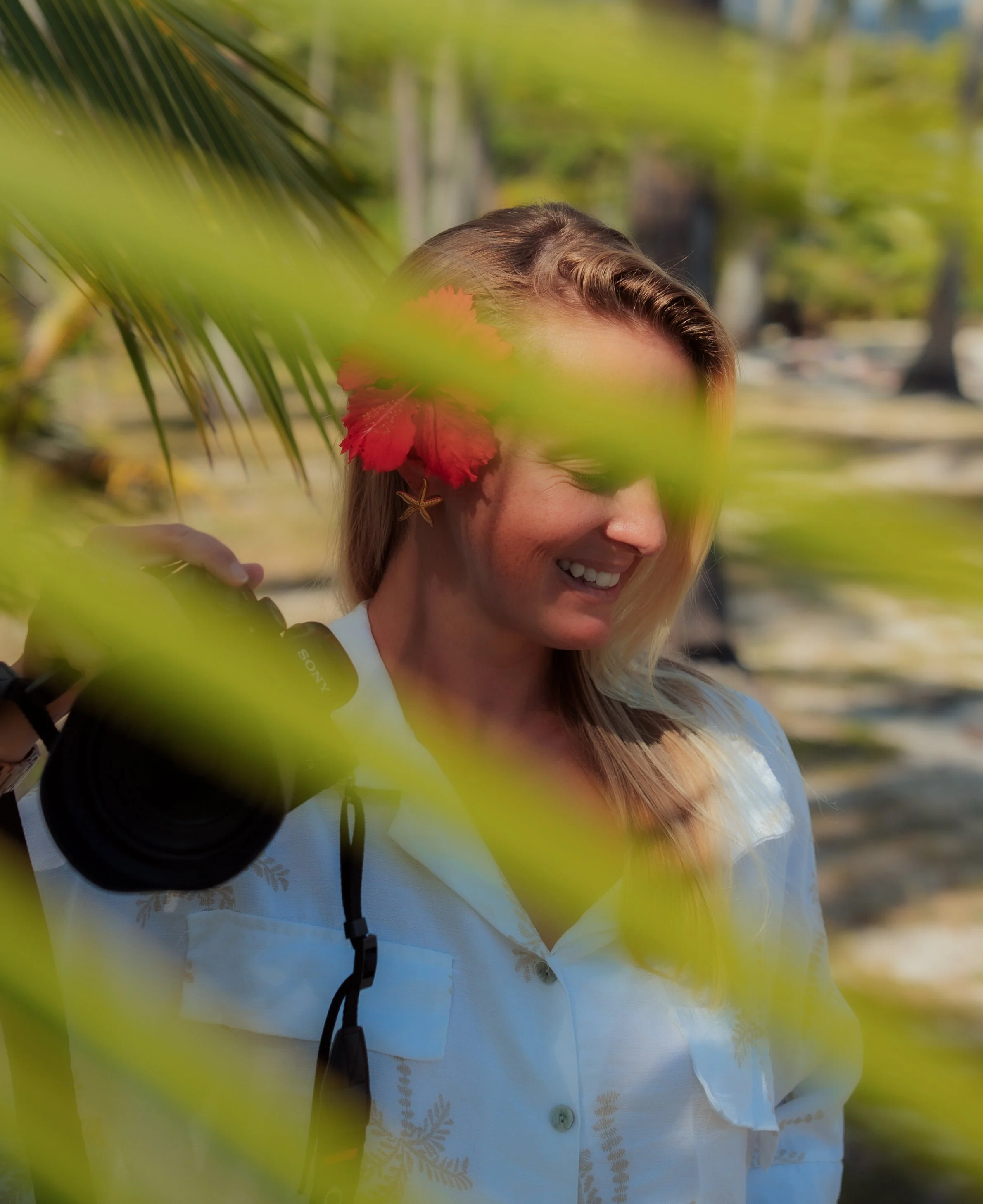 Femme souriante portant une chemise blanche et un sac à dos, partiellement cachée derrière des feuilles de palmier, dans un parc en plein air. Photographe en Polynésie française
