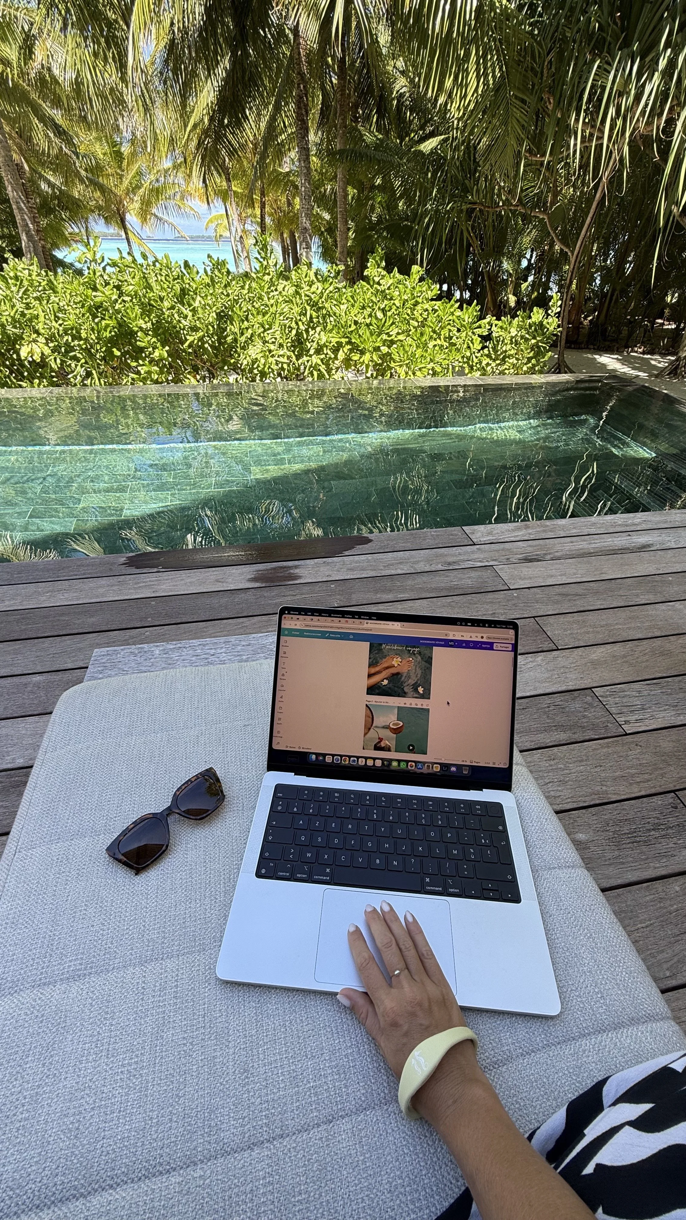 Une personne travaillant sur un ordinateur portable sur une table à côté d'une piscine, avec des lunettes de soleil posées à côté, dans un environnement tropical avec beaucoup de verdure et des palmiers en arrière-plan.