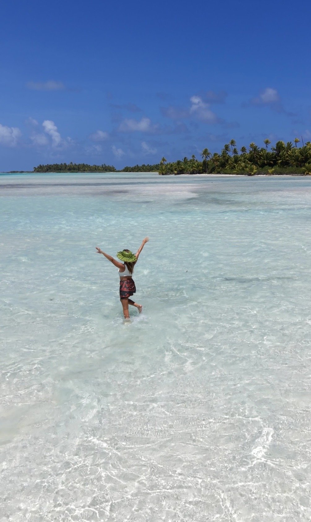 Une jeune fille en maillot de bain et chapeau de paille joue dans l'eau claire d'une plage tropicale avec des cocotiers et un ciel bleu en arrière-plan.