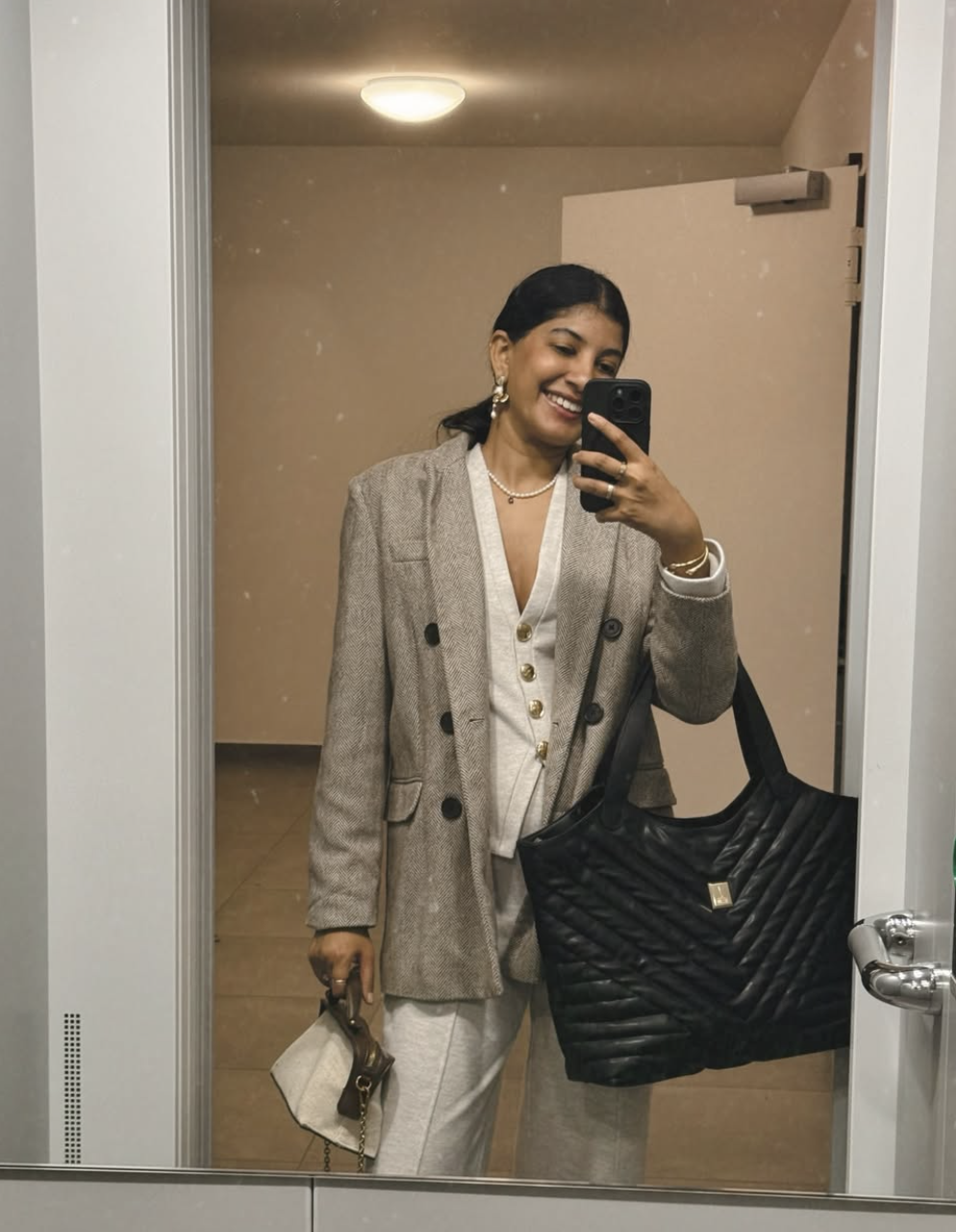 Femme avec boucle d'oreilles, collier de perles, blazer beige, chemise blanche, pantalon beige, tenant un grand sac noir et une petite pochette beige, dans une salle en arrière-plan.