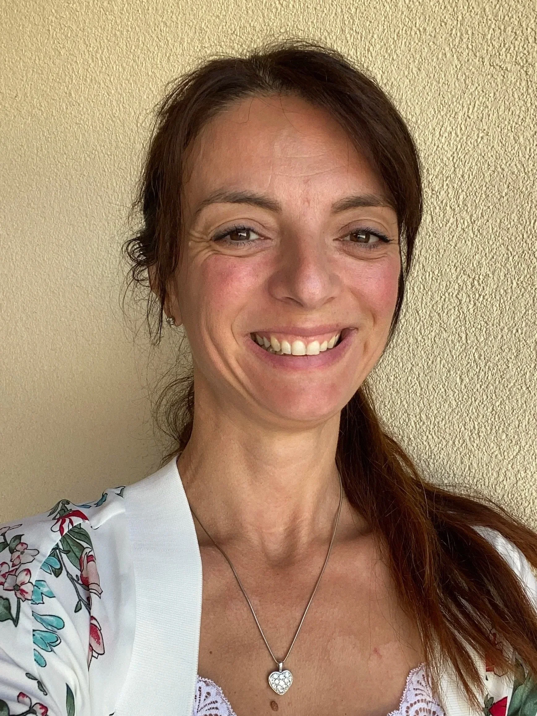 Portrait d'une femme souriante avec des cheveux bruns et une tenue blanche avec des motifs floraux, portant un collier en forme de cœur.