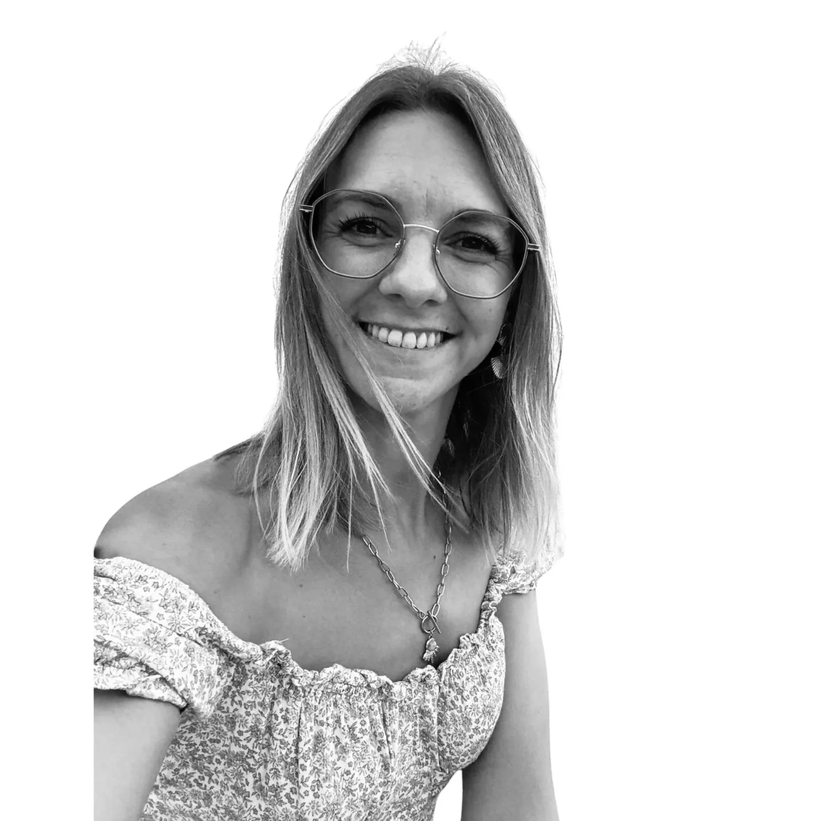 Photo en noir et blanc d'une femme souriante portant de grandes lunettes rondes, une chaîne en or et un haut à épaules dénudées avec un motif floral.