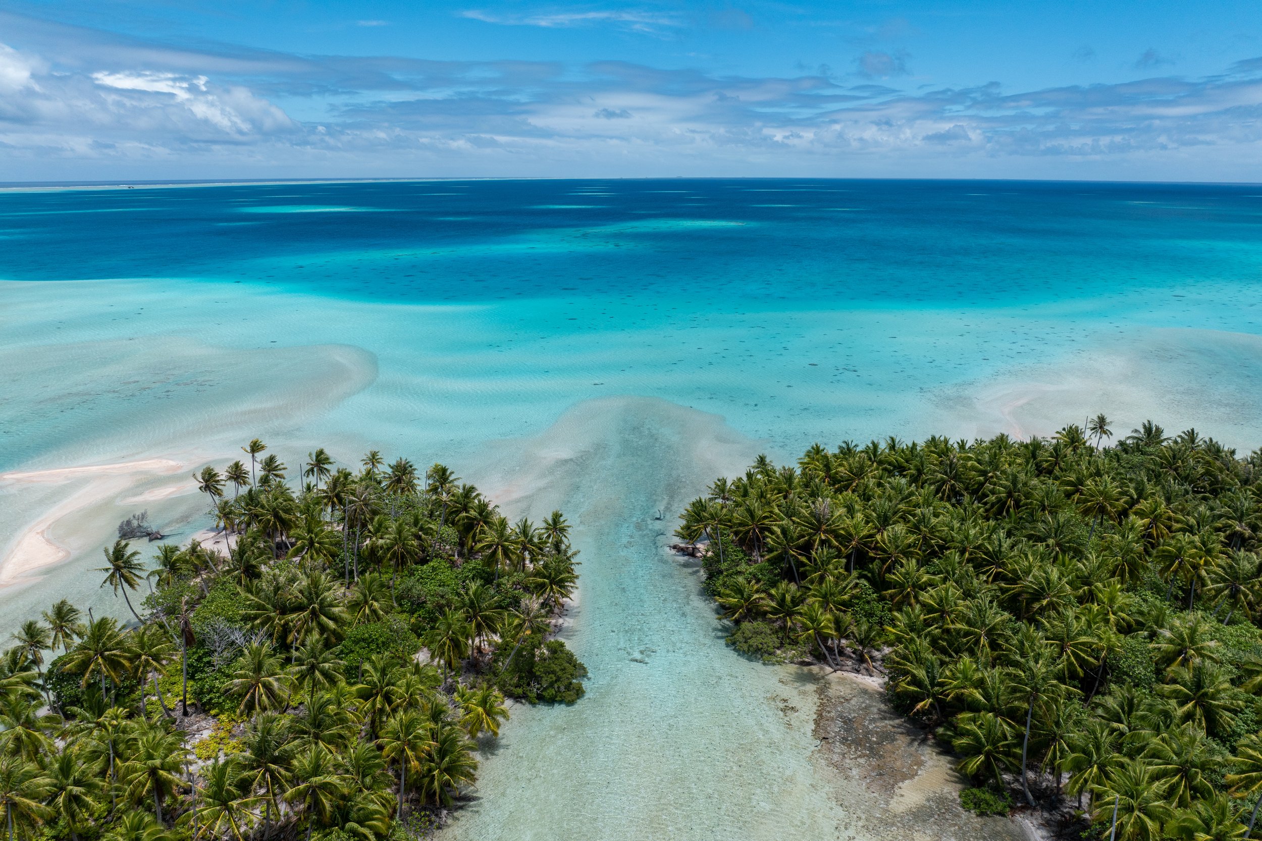 Vue aérienne d'une plage de sable blanc bordée de palmiers, avec une mer turquoise claire et des eaux peu profondes près de la côte, sous un ciel partiellement nuageux.
