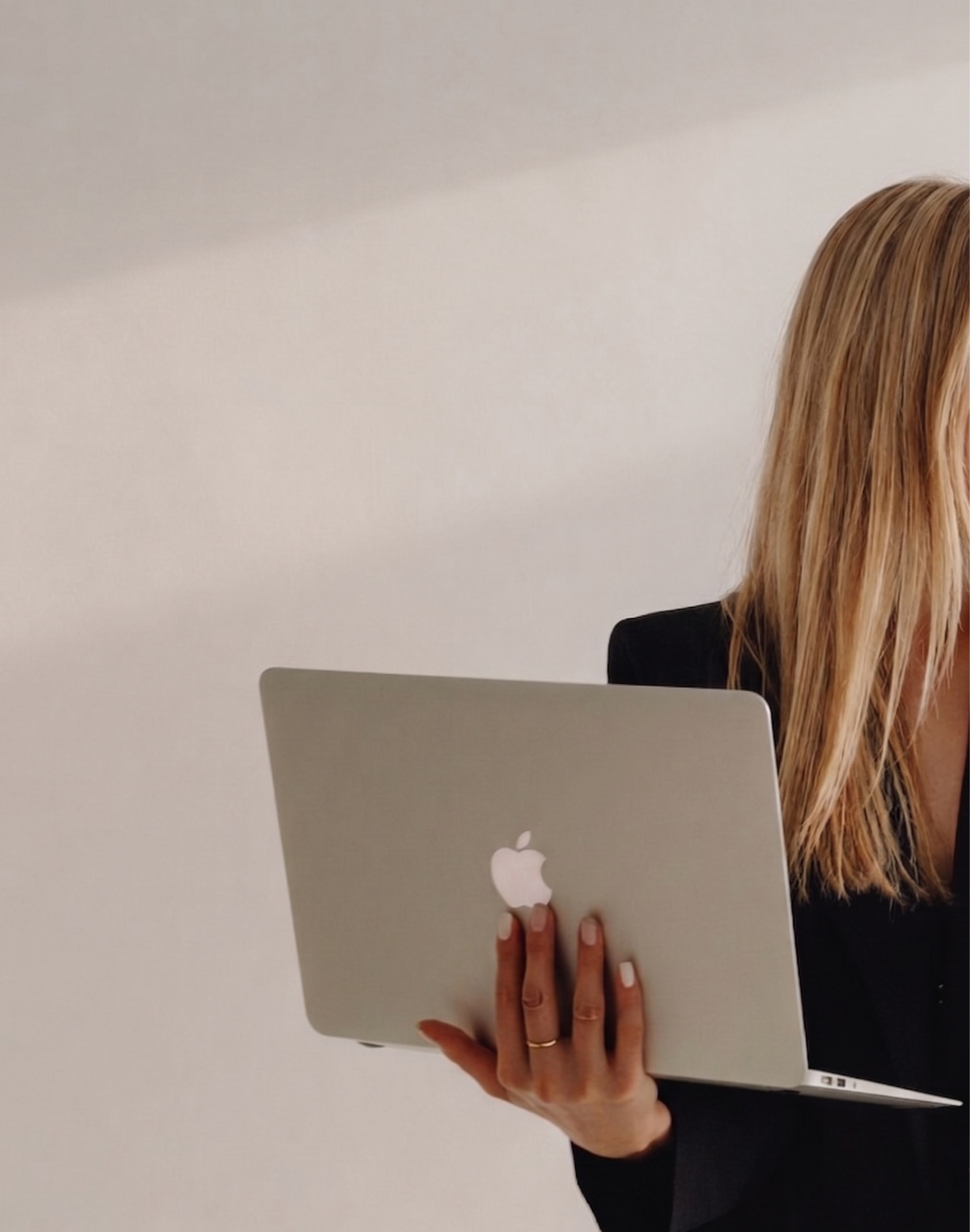 Une personne tenant un ordinateur portable de marque Apple dans la main, avec une main posée sur le clavier. La personne a des cheveux longs, blond foncé, et porte une veste noire. L'image est prise contre un mur blanc.