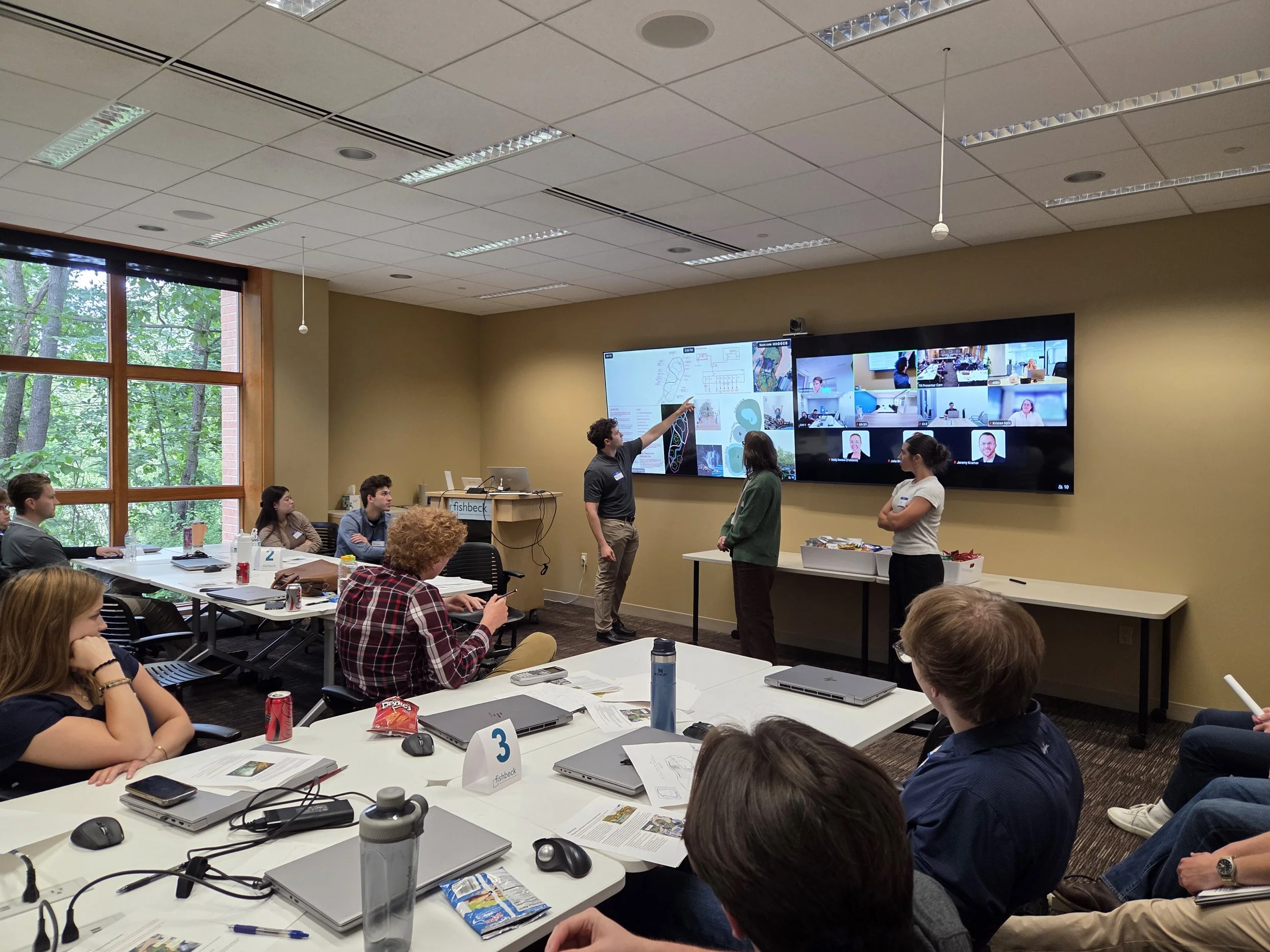 People attending a meeting in a conference room with a presenter pointing at a large digital screen showing graphical information. Attendees are seated at tables with laptops, notebooks, and snacks. The room has large windows with trees outside.