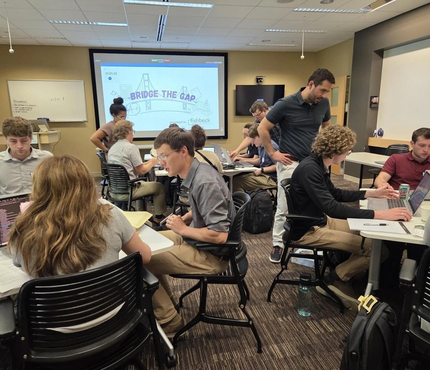 A classroom or workshop setting with students working on laptops and discussions occurring. A large screen displays a presentation titled 'Bridge the Gap.' Some students are engaged in group activities, while others work independently.