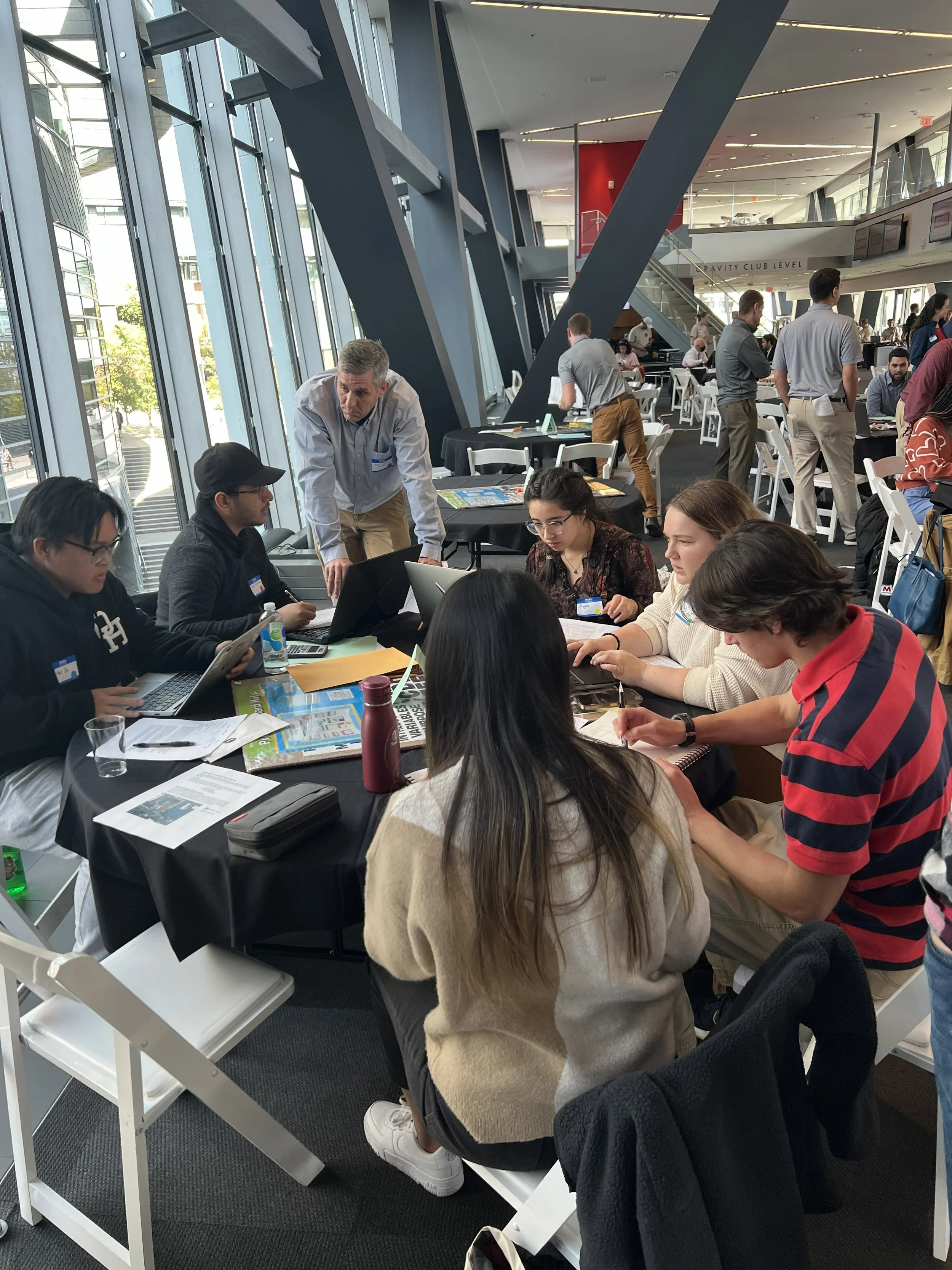 Indoor conference or workshop space with people seated at tables with laptops, papers, and drinks, and some standing or walking around, with large windows and modern architecture in the background.
