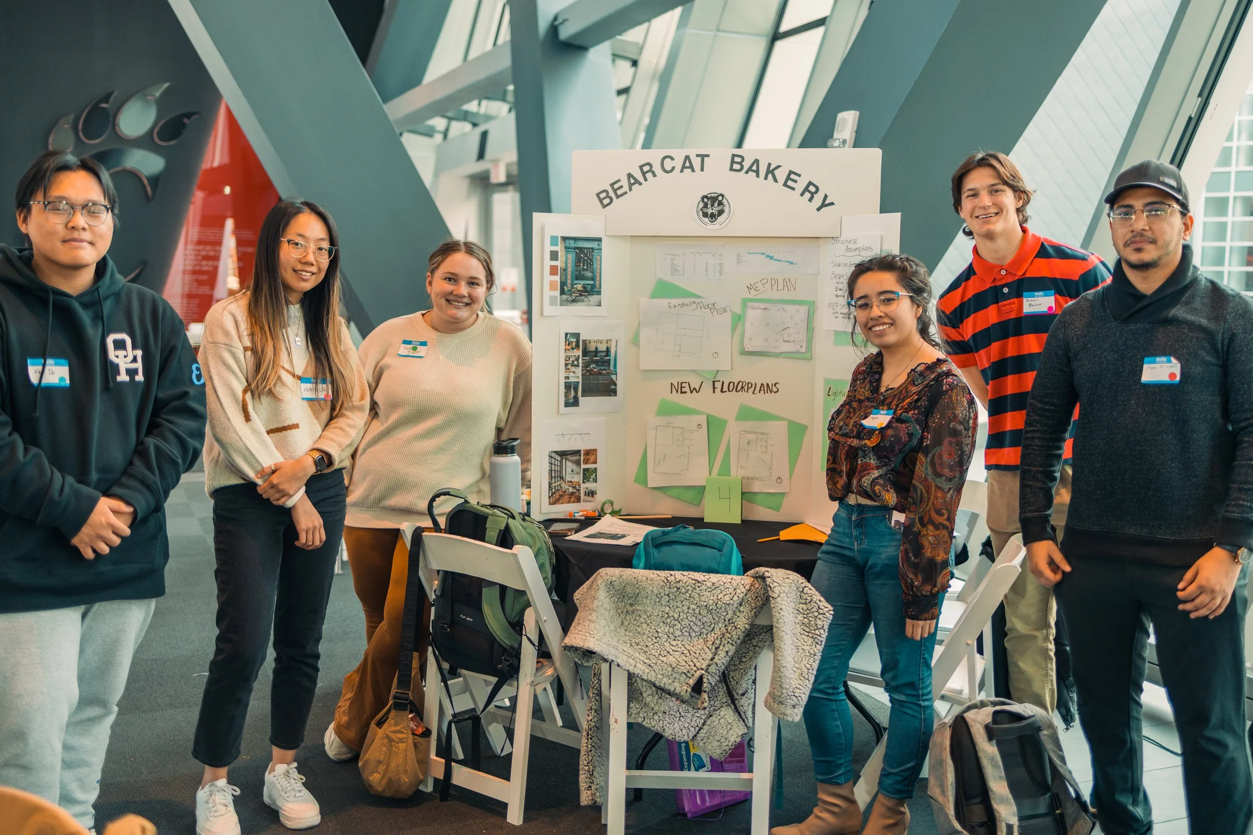 Group of six young people standing in front of a project display booth labeled 'Bearcat Bakery' in a large indoor space with modern architectural design.