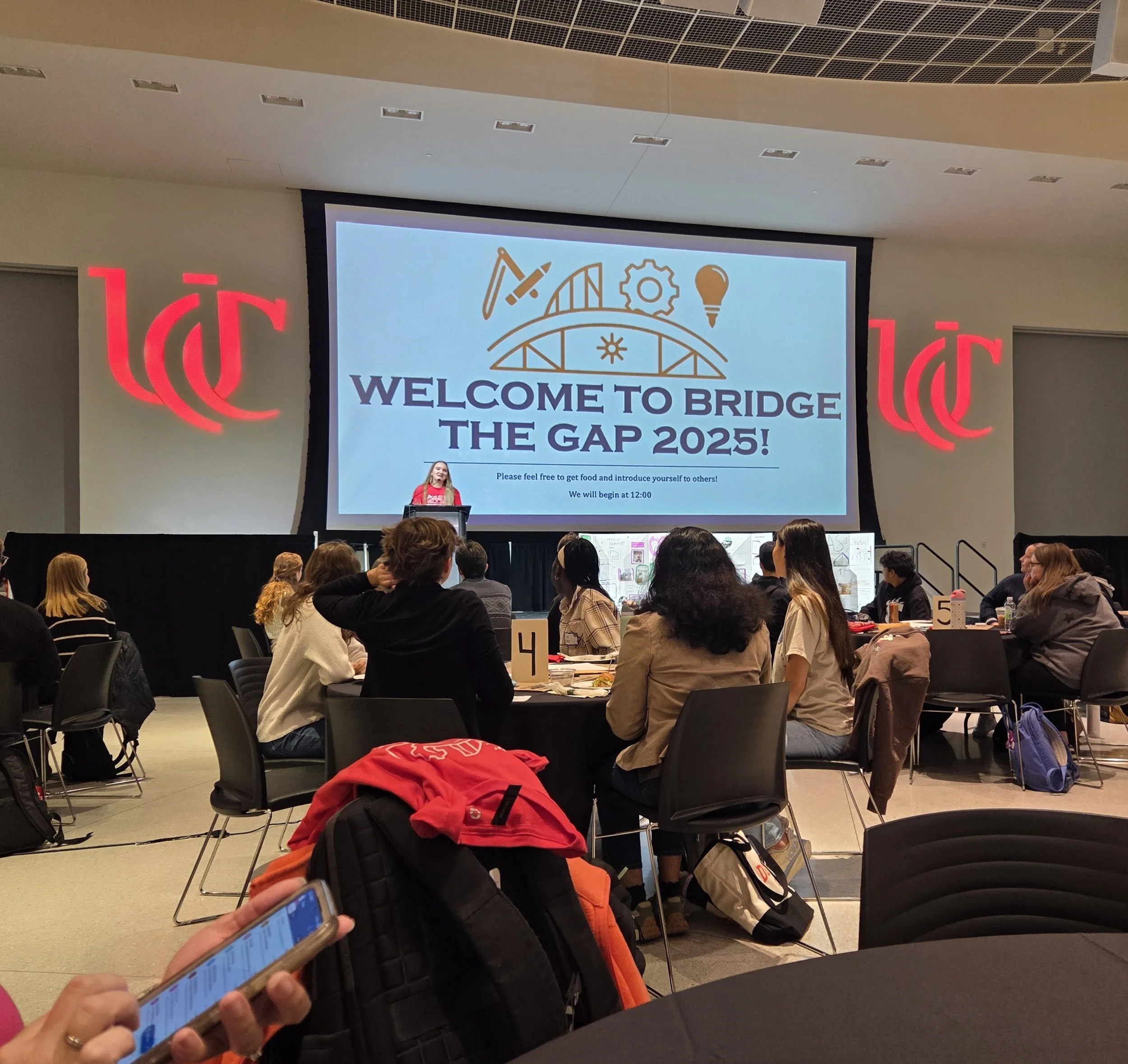 A large indoor event with many people seated at tables, facing a stage with a projector screen that reads 'Welcome to Bridge the Gap 2025!'. A woman with long hair, wearing a red shirt, is standing at a podium on the stage, speaking to the audience. The room has a high ceiling with ventilation panels, and red illuminated walls with a design or logo.