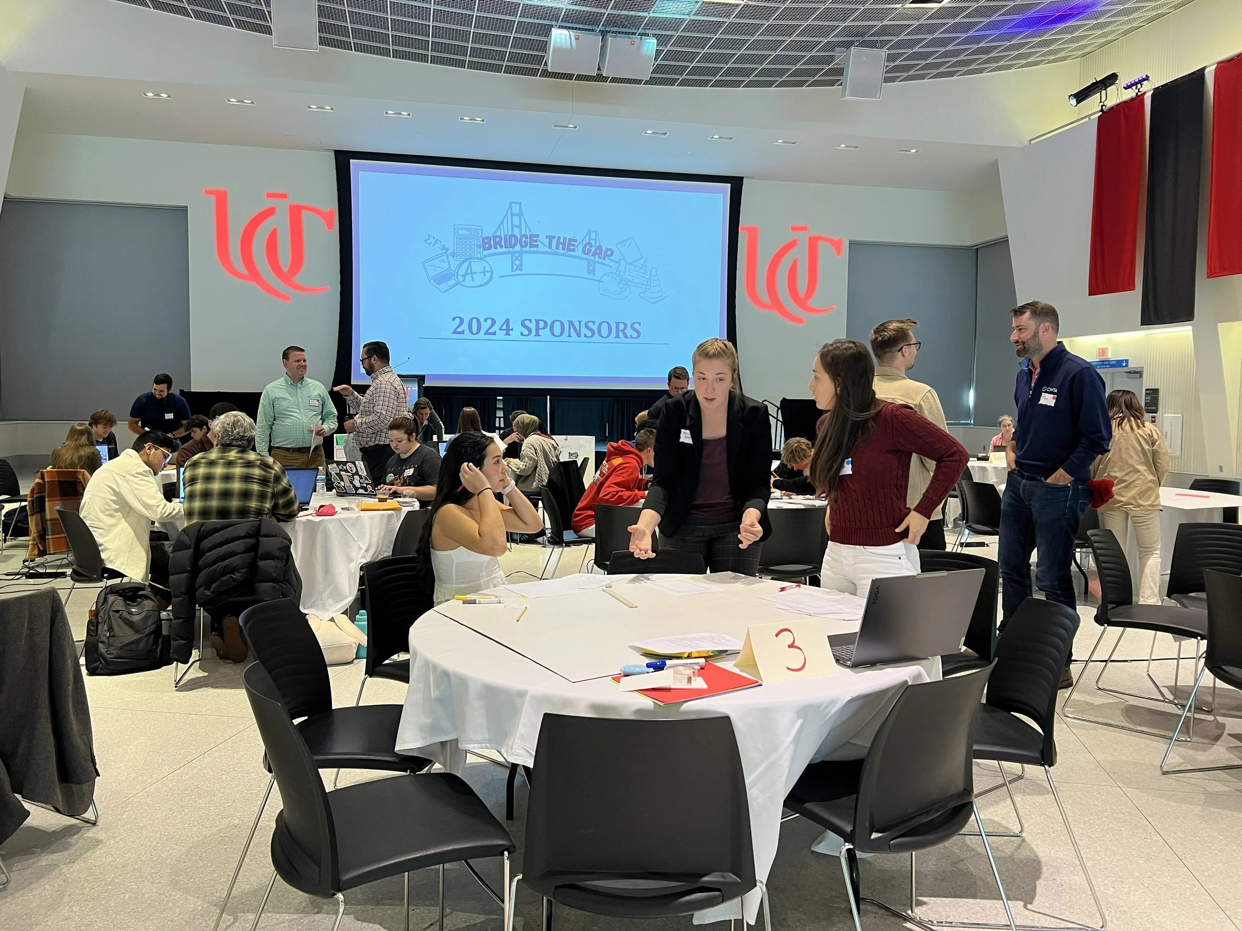 People attending a conference or workshop in a large hall, sitting at round tables with laptops and papers, with a large screen displaying 'Bridge the Gap' and '2024 Sponsors'. Several individuals are standing and talking, and there are large red and black banners hanging from the ceiling.