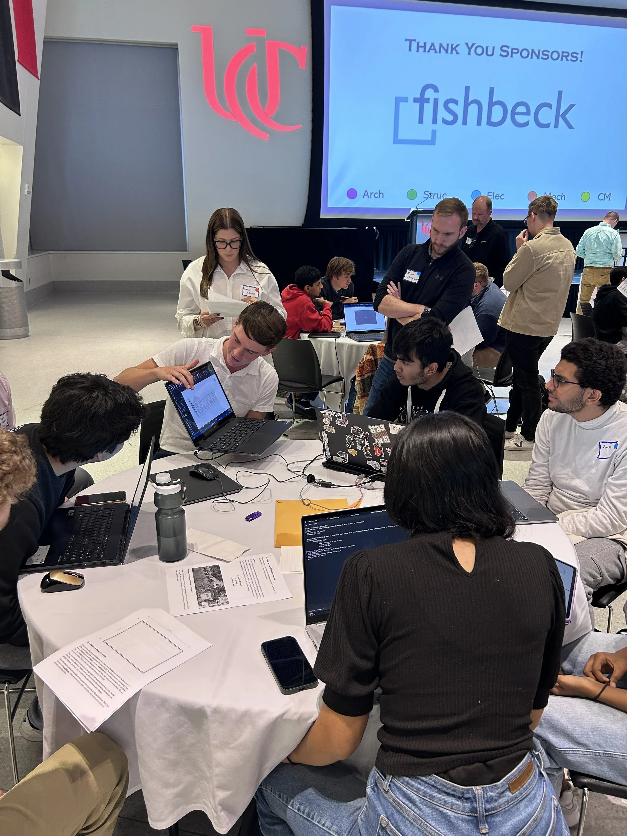 People gathered around a table working on laptops and reading papers at a conference or workshop, with a large screen in the background displaying a thank you message to sponsors and a logo.