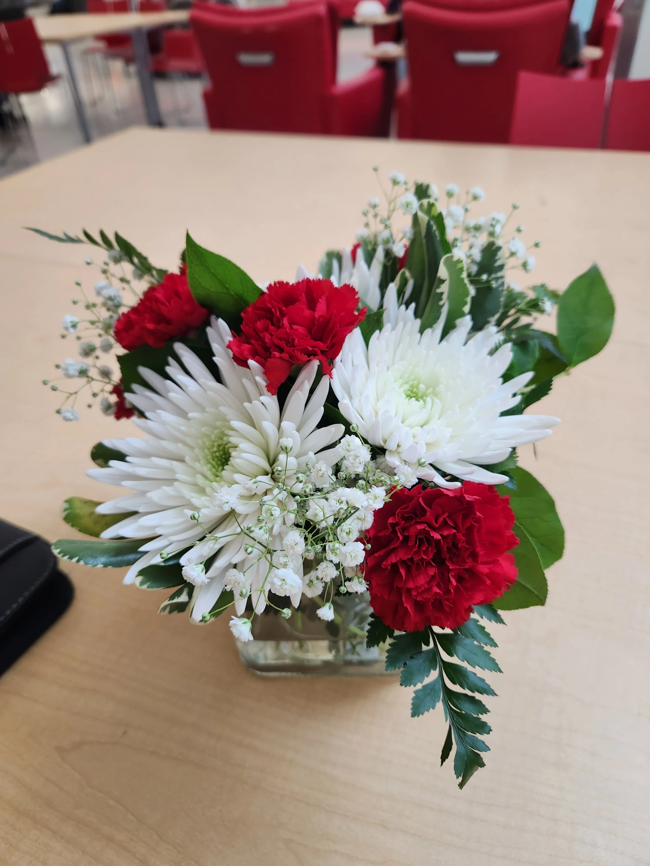 A small bouquet of white and red flowers and foliage on a light wooden table in an indoor setting with red chairs in the background.