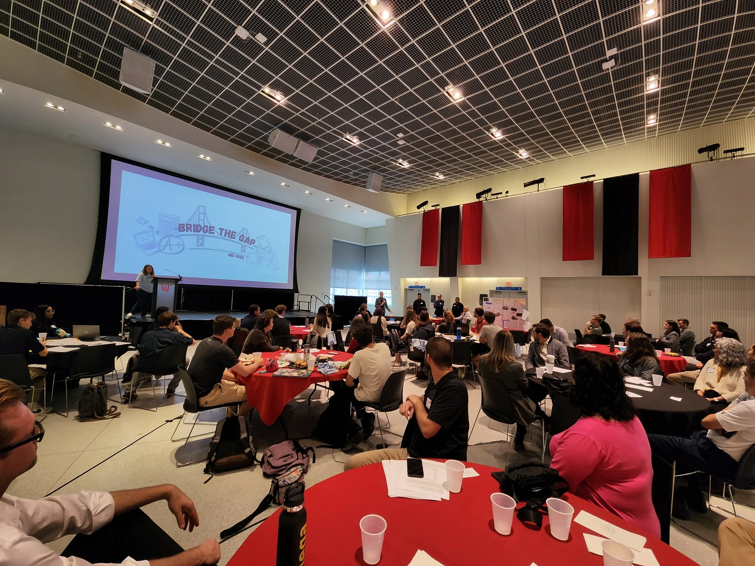 A large conference room filled with people sitting at round tables covered with red tablecloths, attending a presentation. A woman stands at a podium near a large screen displaying a slide with the title 'Bridge the Gap' and an illustration of a bridge and a cityscape. The room has a high ceiling with a grid pattern, and walls decorated with red and black vertical banners.