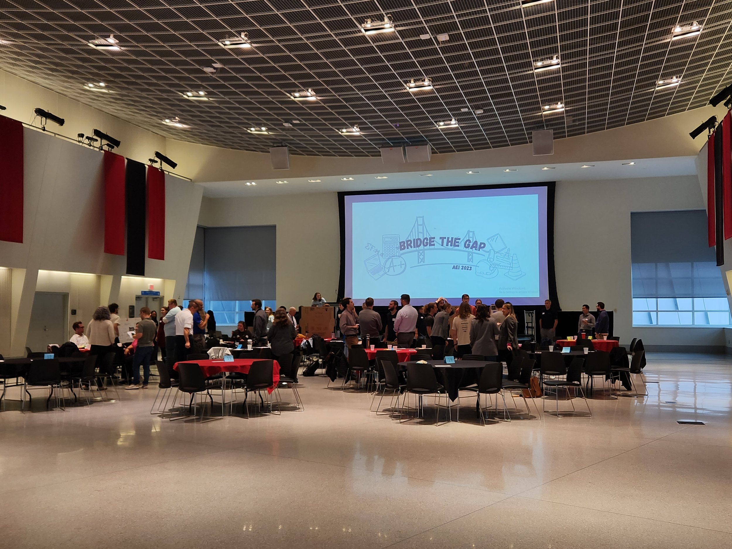 An indoor conference or event space with a high, grid-patterned ceiling, red and black curtains, a large projection screen displaying a slide titled "Bridge the Gap" with illustrations of building tools and a bridge, and people gathered around tables and standing in small groups.