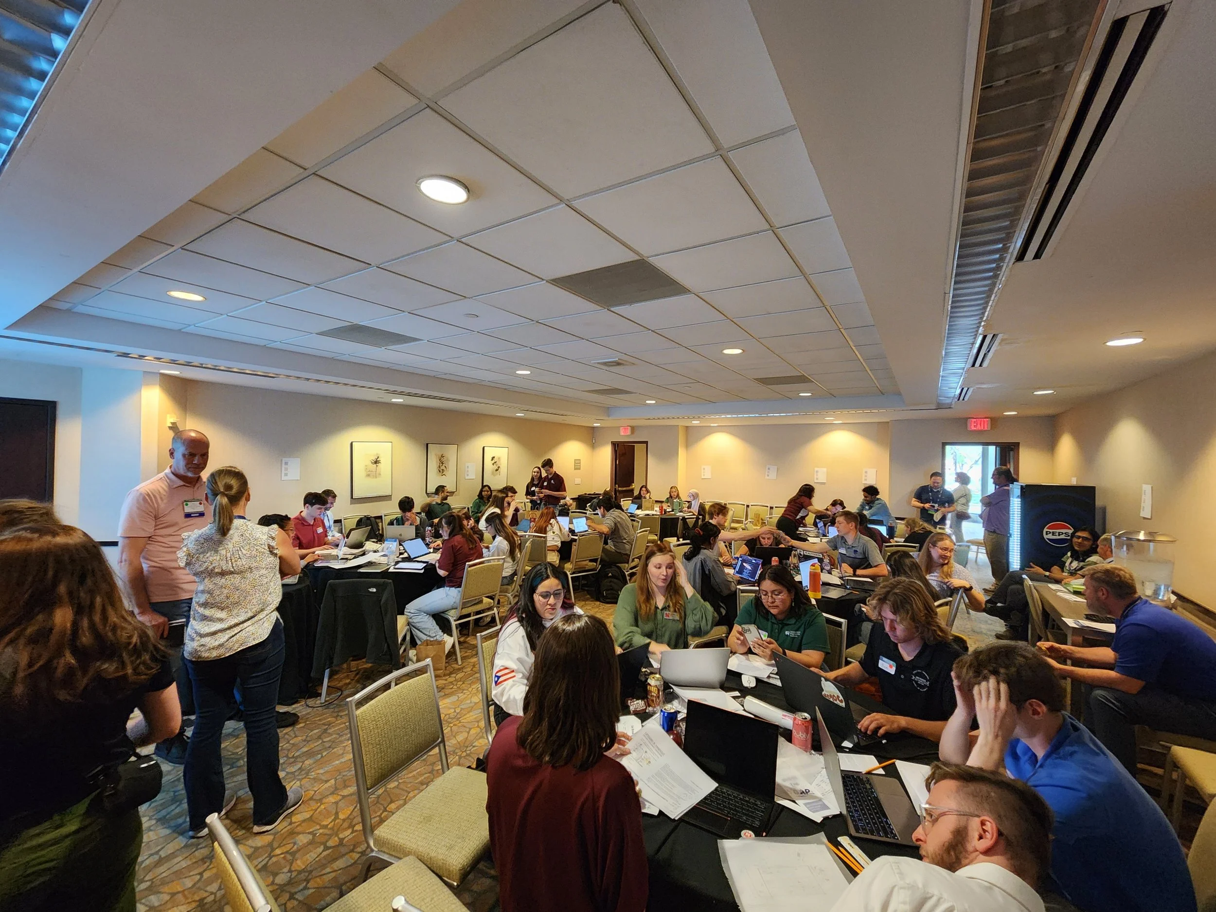 A busy conference room filled with people working on laptops and papers, with some standing and socializing. The ceiling has recessed lights and air vents, and there are framed pictures on the walls. The room has a carpeted floor and appears to be part of a professional or academic event.