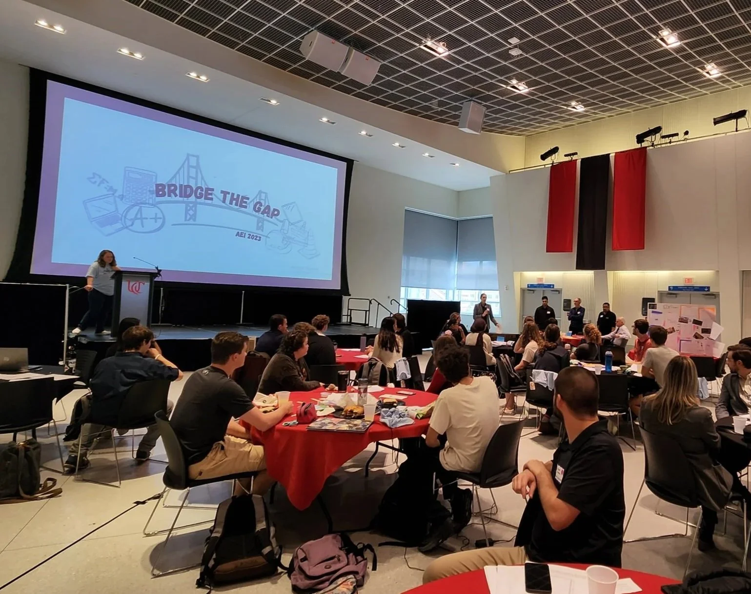 A large conference room filled with people sitting at round tables covered with red tablecloths, attending a presentation. A large screen displays a graphic of a bridge and the words "Bridge the Gap". Several individuals stand at the front of the room near the screen, speaking or presenting. The room has high ceilings with a grid design, and two large banners, one red and one black, hang on the wall behind the presenters.