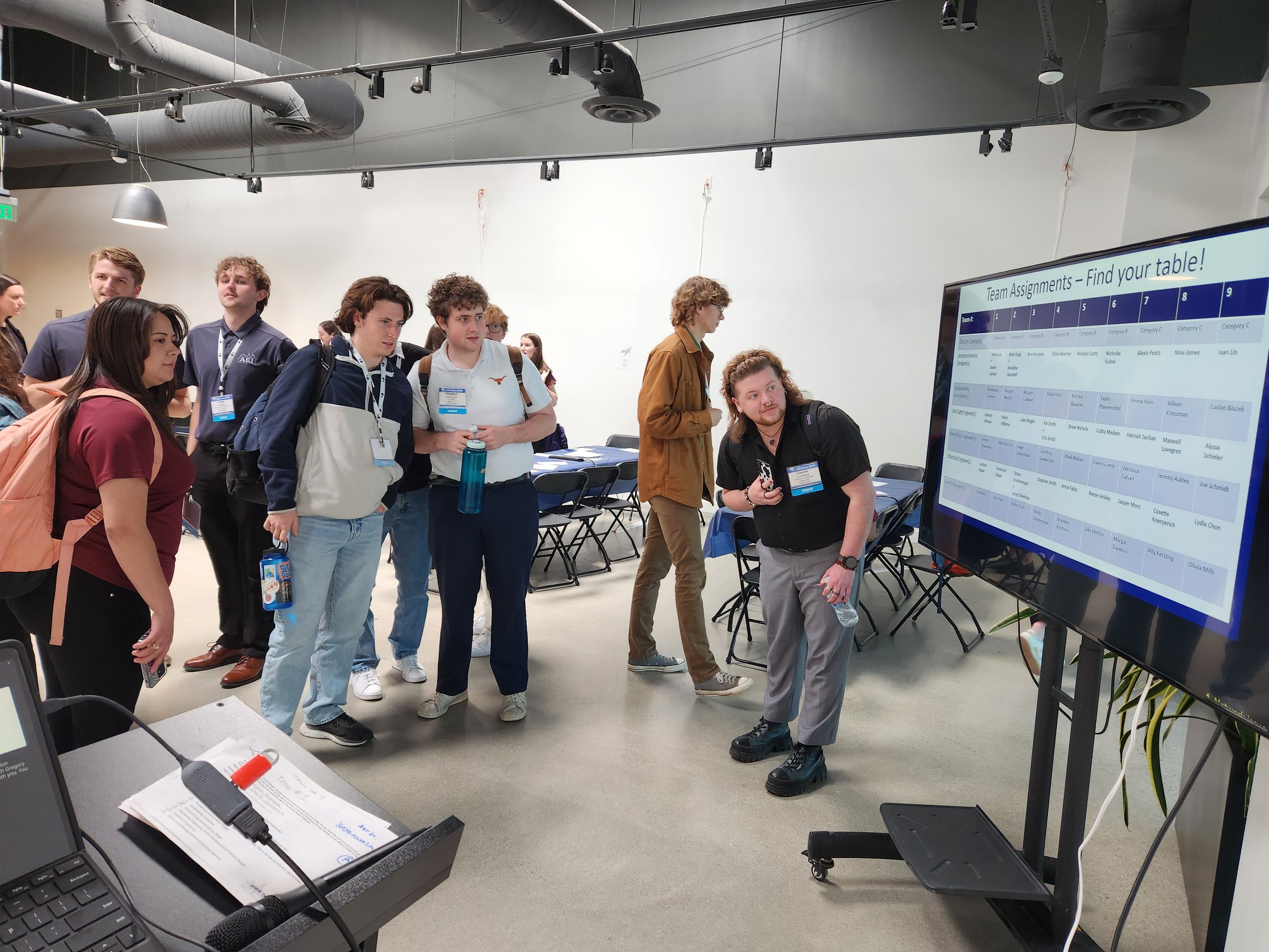 Group of young adults standing in a line, looking at a large digital screen displaying team assignments at a conference or workshop.