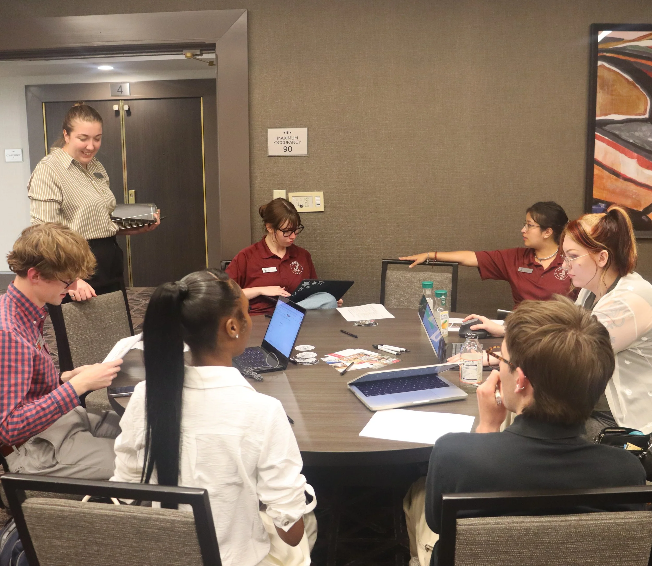 A group of people seated around a conference table, some using laptops, in a meeting room with a woman standing and speaking, and a sign on the wall indicating a maximum occupancy of 90.
