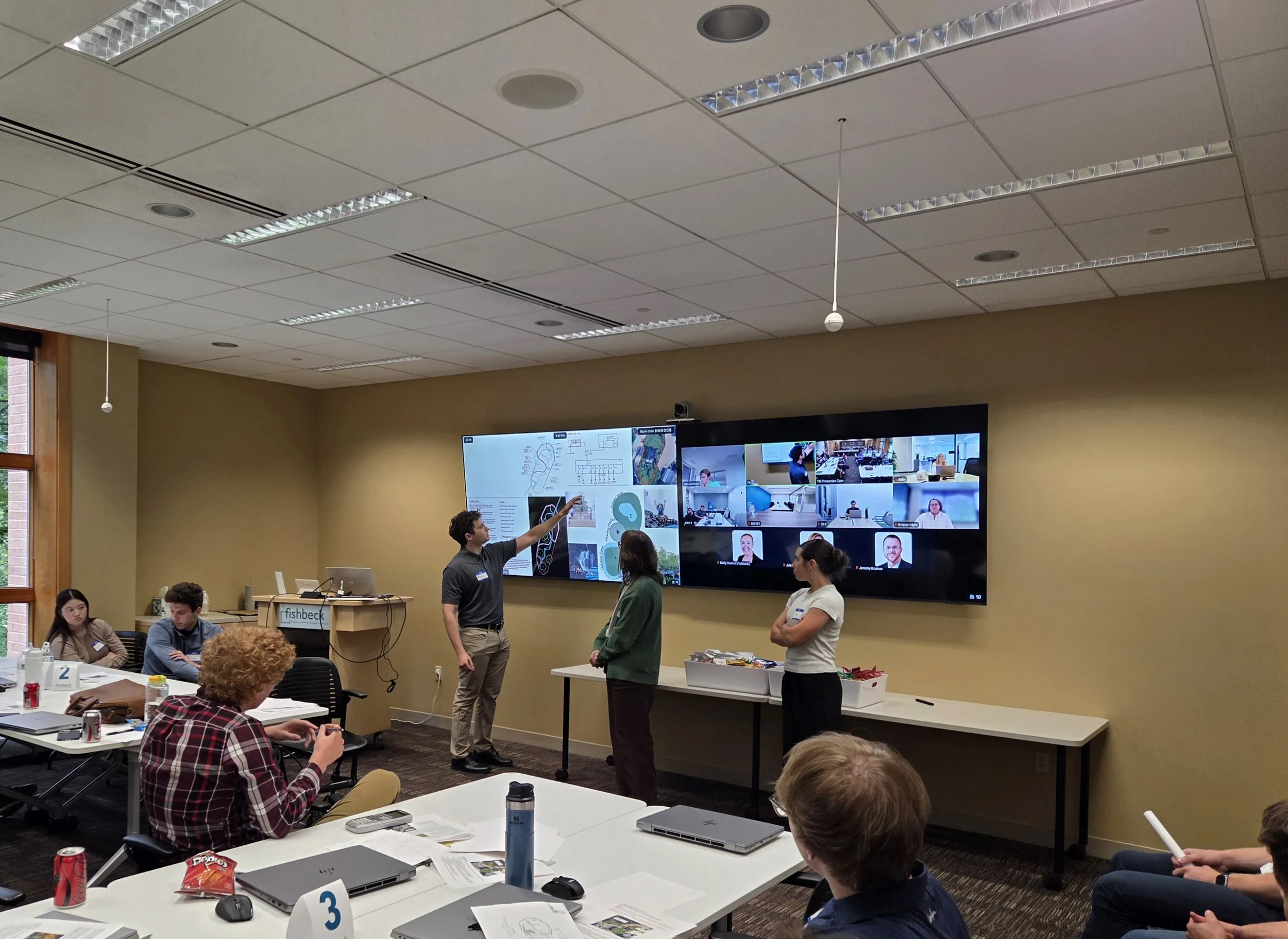 A classroom or conference room with people sitting at tables and a person standing and pointing at a large display screen showing a video conference with multiple participants.