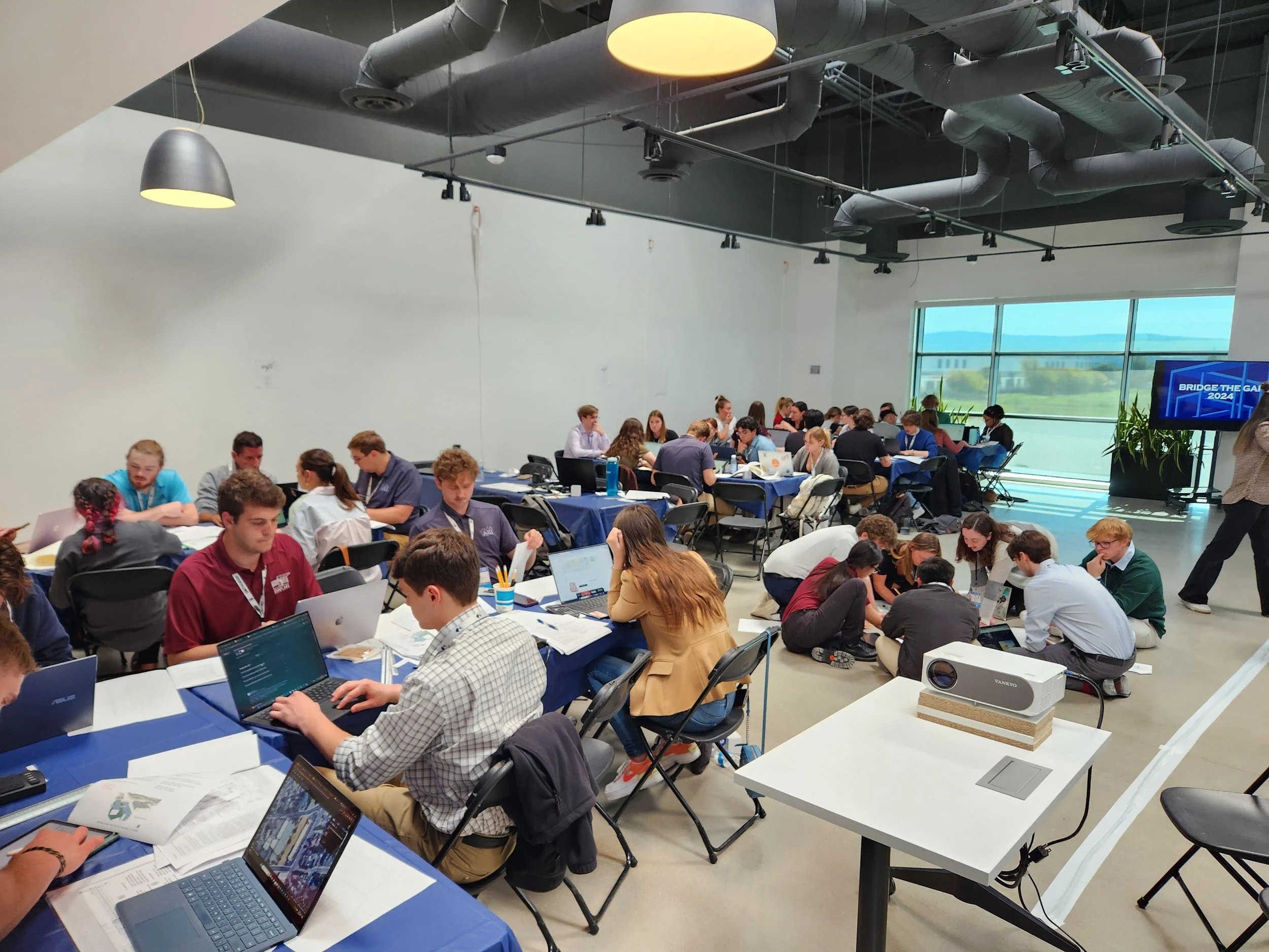 A large group of people working on laptops and discussing around tables in a modern conference room with high ceilings, large windows, and an industrial design. There is a screen displaying a presentation titled 'Bridge the Gap 2024' and a projector on a white table.