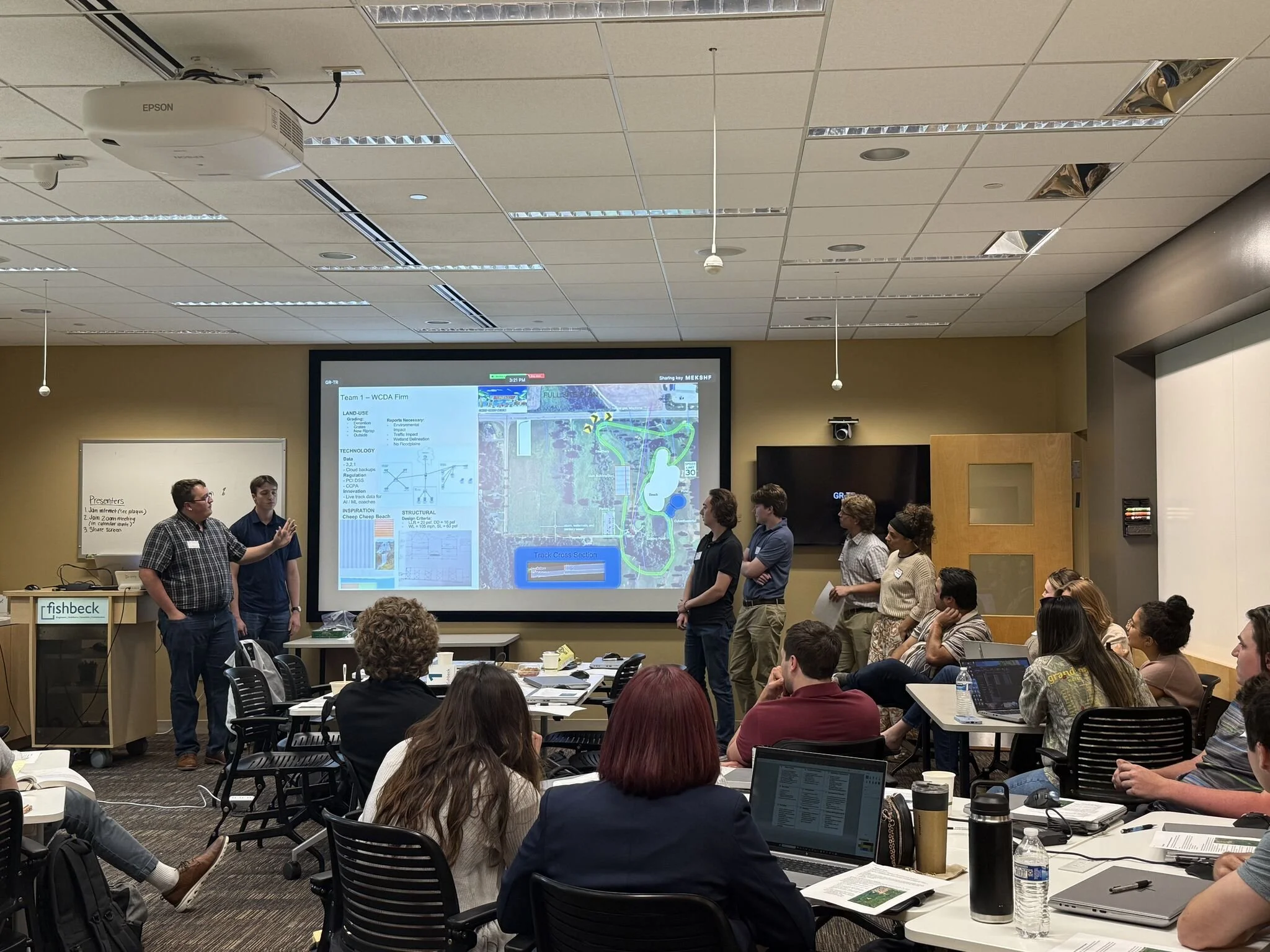 A conference room filled with people attending a presentation. A group of presenters are standing in front of a large screen displaying a map and text. The room has some ceiling tiles missing, and a projector is mounted on the ceiling.