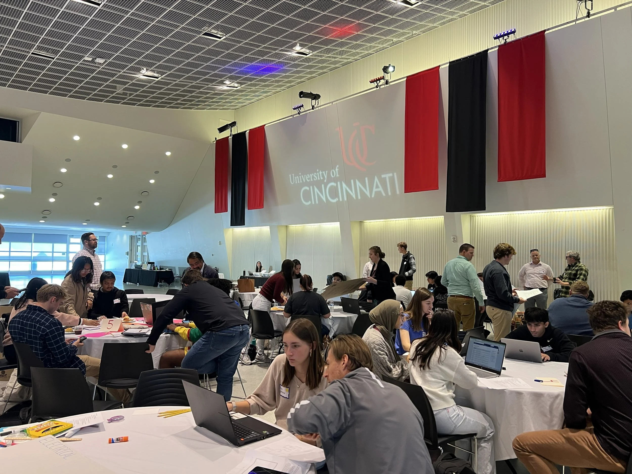 People working together at tables in a large, well-lit conference room at the University of Cincinnati, with a projection screen displaying the university's name and red and black banners hanging from the ceiling.