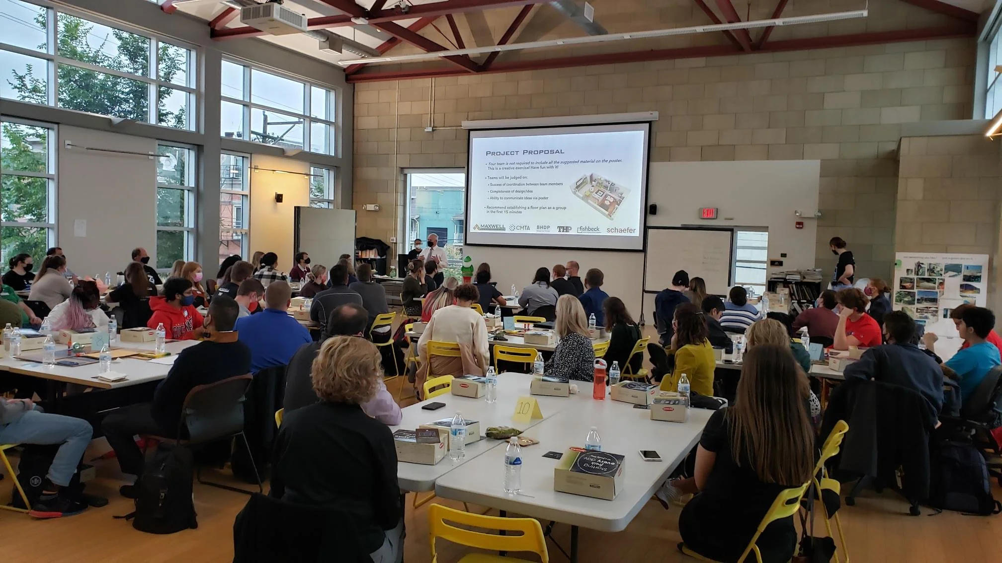 A large group of people attending a conference in a spacious room with high ceilings and large windows. There is a speaker at the front near a projection screen displaying a presentation titled 'Project Proposal'. Attendees are seated at tables with notebooks, water bottles, and presentation materials, listening to the speaker.