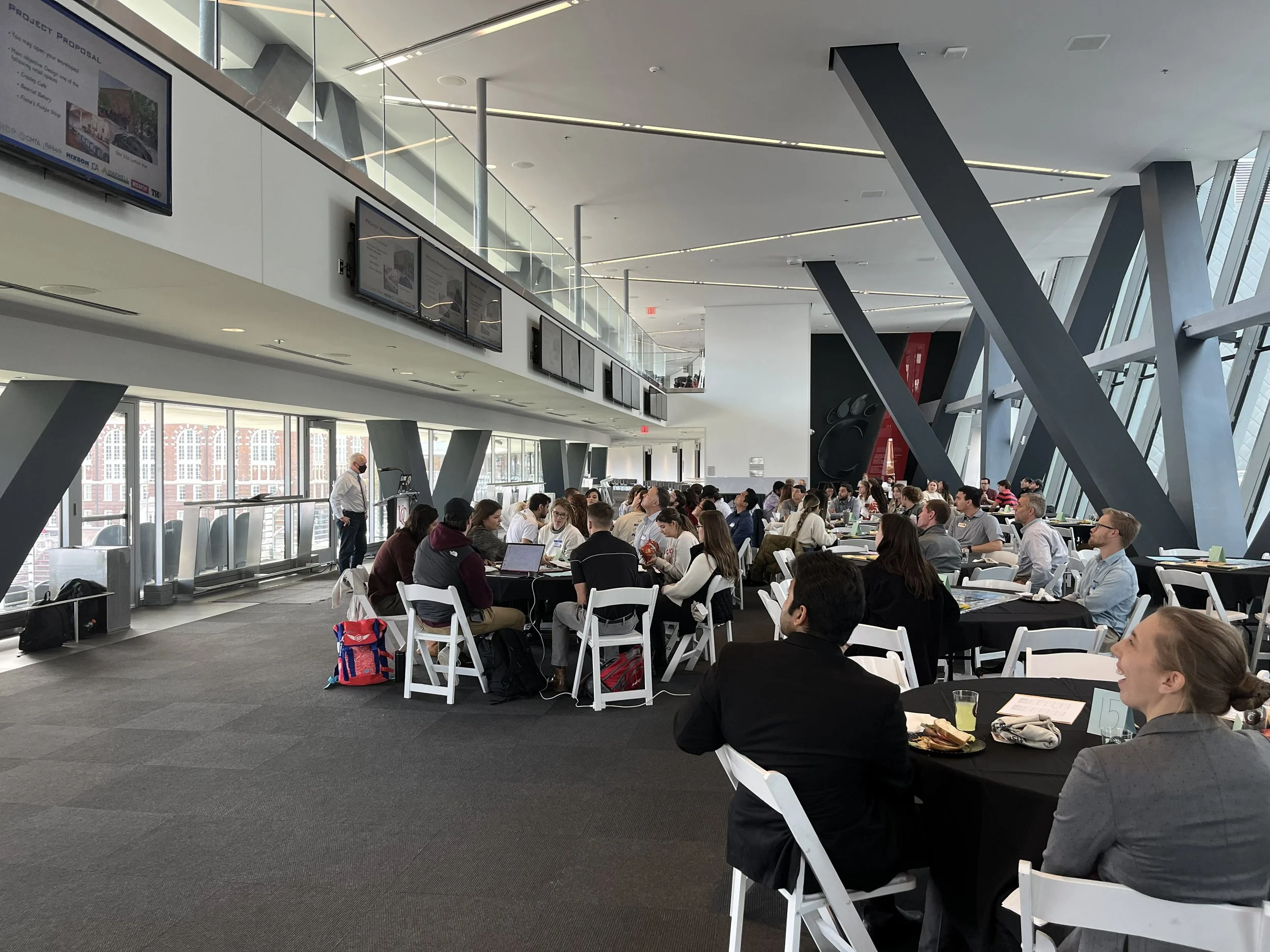 A large, bright conference center with people seated at tables and some standing, having meals or working on laptops. There are large windows with a view of the city outside, digital display screens on the upper level, and modern architectural elements such as big angled supports and glass railings.