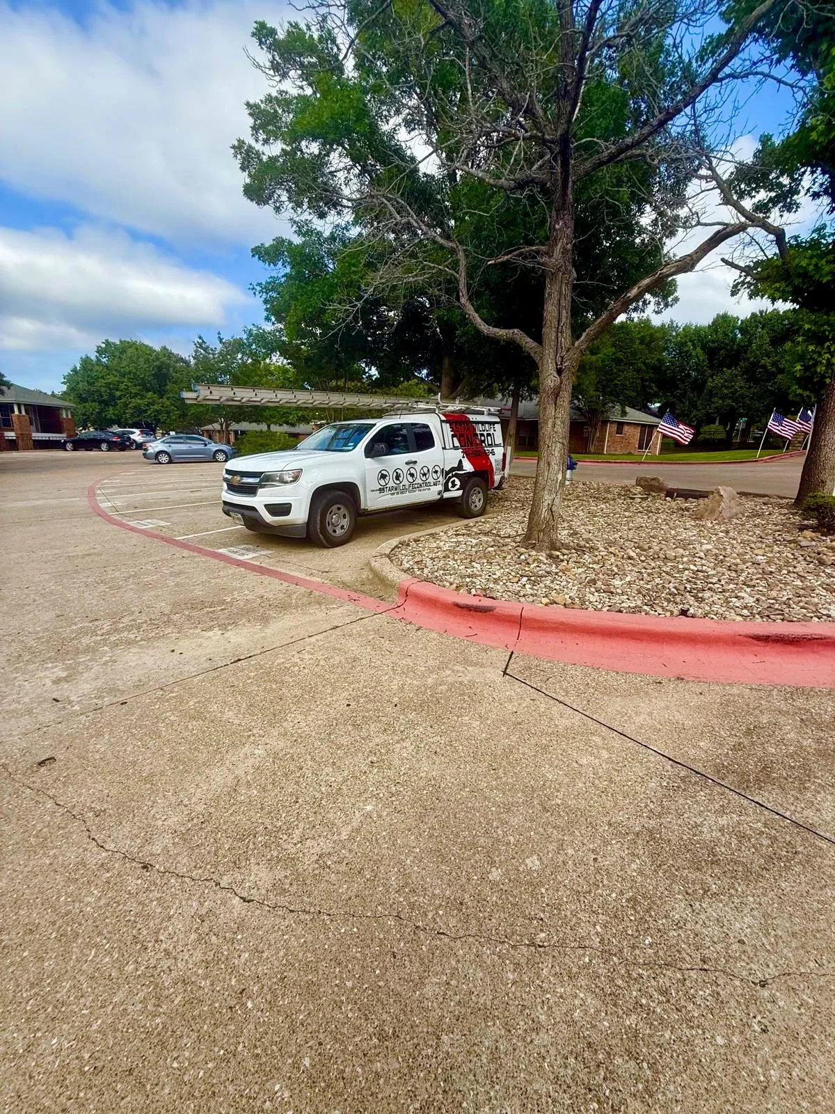 A wildlife removal and control work truck at job site with a ladder on top parked in a mostly empty parking lot. There are trees, American flags, and a brick building in the background. 