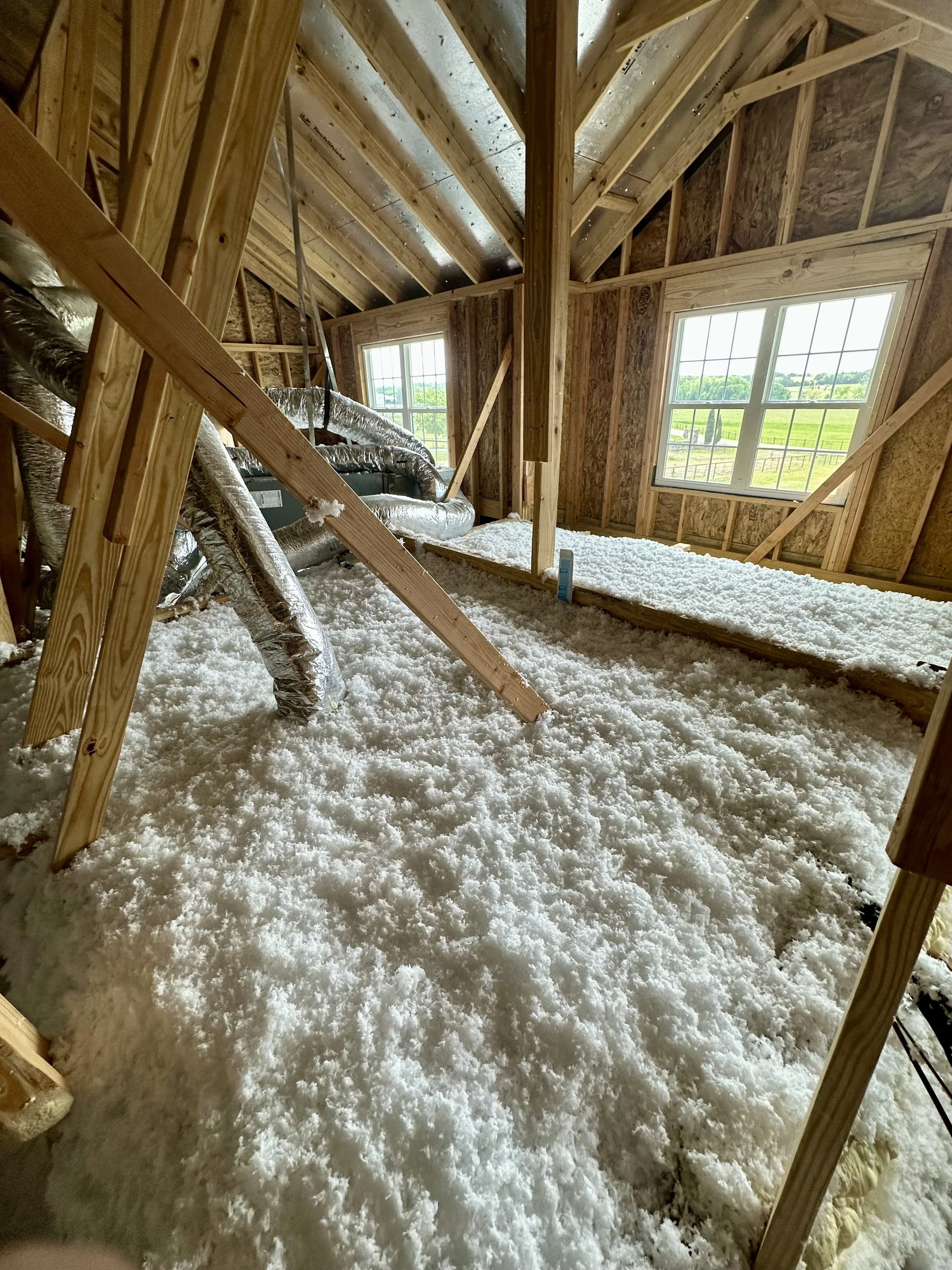 Interior of a house under construction, showing exposed wood framing, a sloped roof with insulation, and a pile of white insulation material on the floor near a window with a scenic view outside.