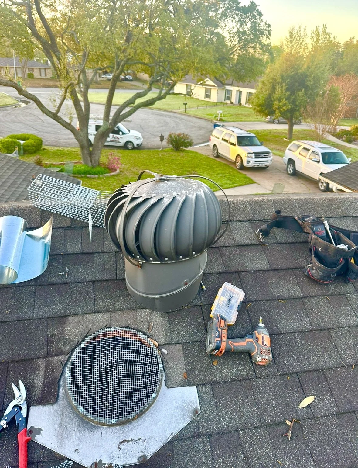 View from a rooftop showing tools, parts of a vent cover, and a vent pipe on a shingled roof. In the background, there are parked cars, a tree, a grassy area, and houses.