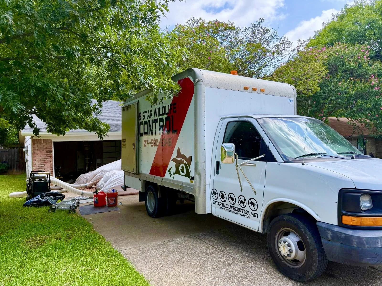 A wildlife control truck parked on a residential driveway with a green and white house and trees in the background. work truck parked outside insulation removal job site