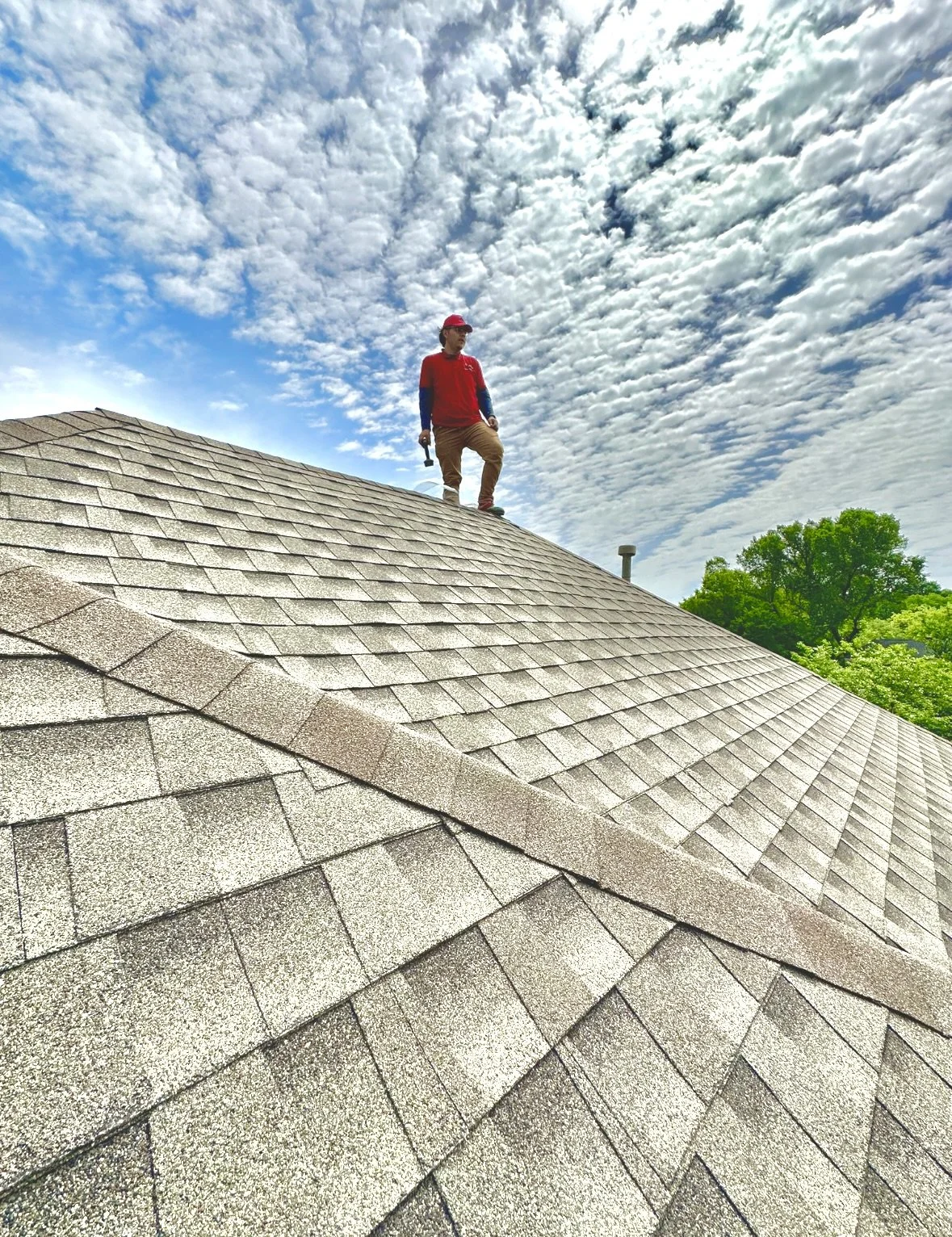 A person in a red shirt and tan pants is standing on the peak of a shingled roof, holding a tool in their right hand, with trees and a cloudy sky in the background. inspection roof for rat, squirrel, raccoon, possum