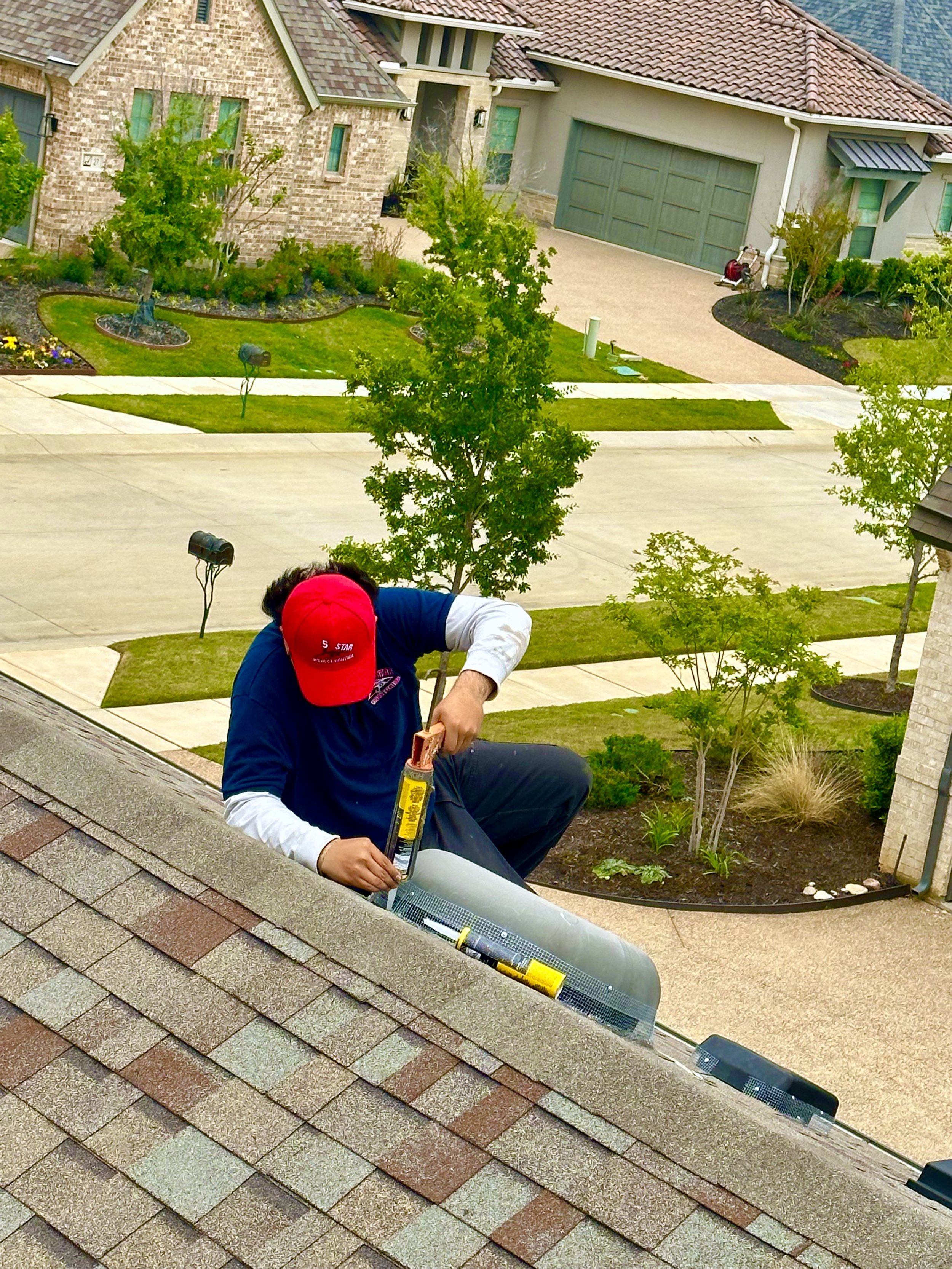 A wildlife control and removal company on a roof with shingles, using a caulking gun to seal around a vent pipe. The person is wearing a red cap and a blue jacket and is kneeling on the roof.