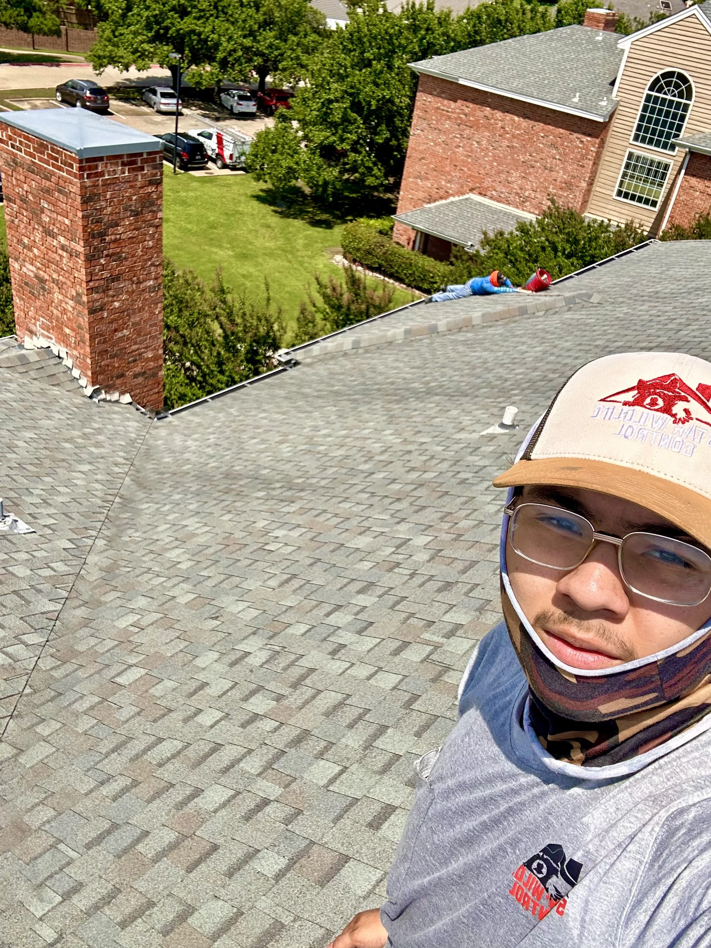 A person wearing glasses, a baseball cap, and a face mask with a logo, taking a selfie on a house rooftop with a suburban neighborhood in the background, including trees, parked cars, and houses. inspection roof