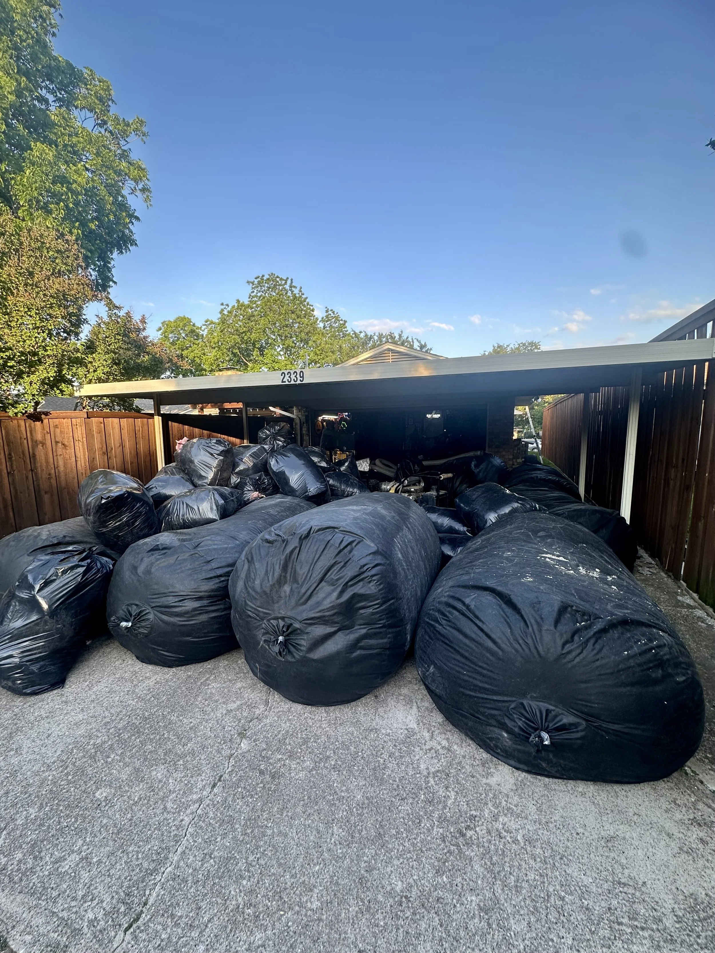 Numerous black garbage bags filled with trash are piled on the driveway next to a wooden fence and backyard, with a carport structure overhead and trees in the background under a clear blue sky. all bags filled with old insulation. insulation removal
