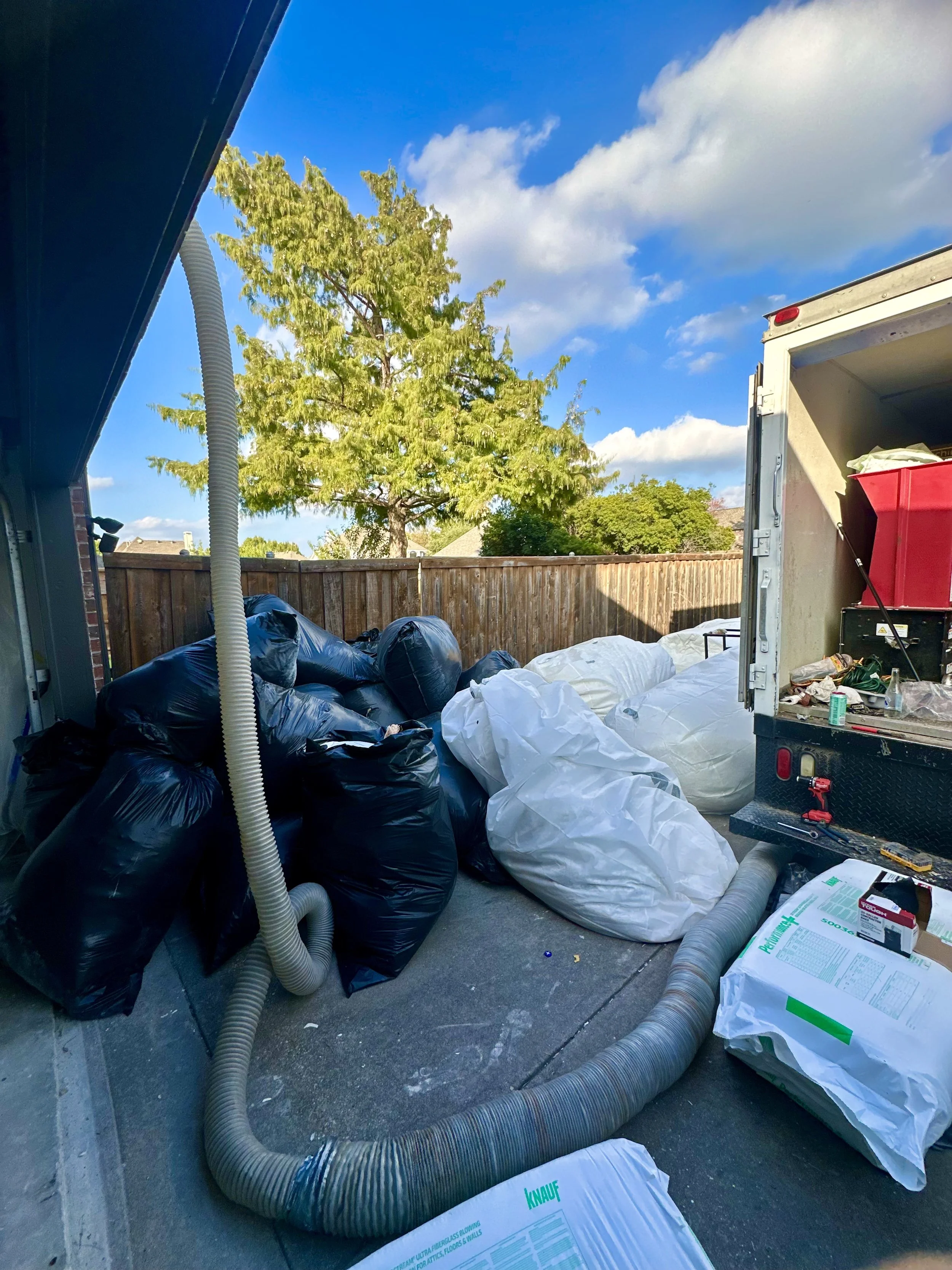 Open truck filled with various bags and construction materials, with large flexible hoses and boxes around it, set in a backyard with a tree, wooden fence, and blue sky with white clouds. insulation install and removal