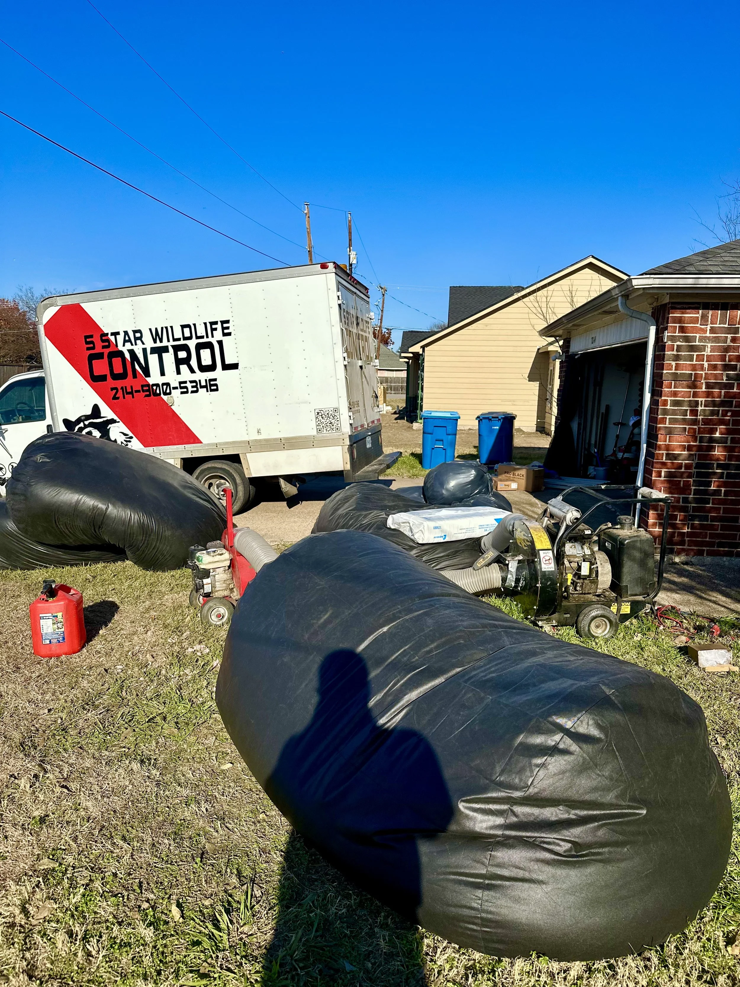 Yard with equipment, black bags, a white truck with '5 Star Wildlife Control' on the side, a small generator, blue trash cans, and a garage with an open door, under a clear blue sky. this company is performing a insulation removal and install service