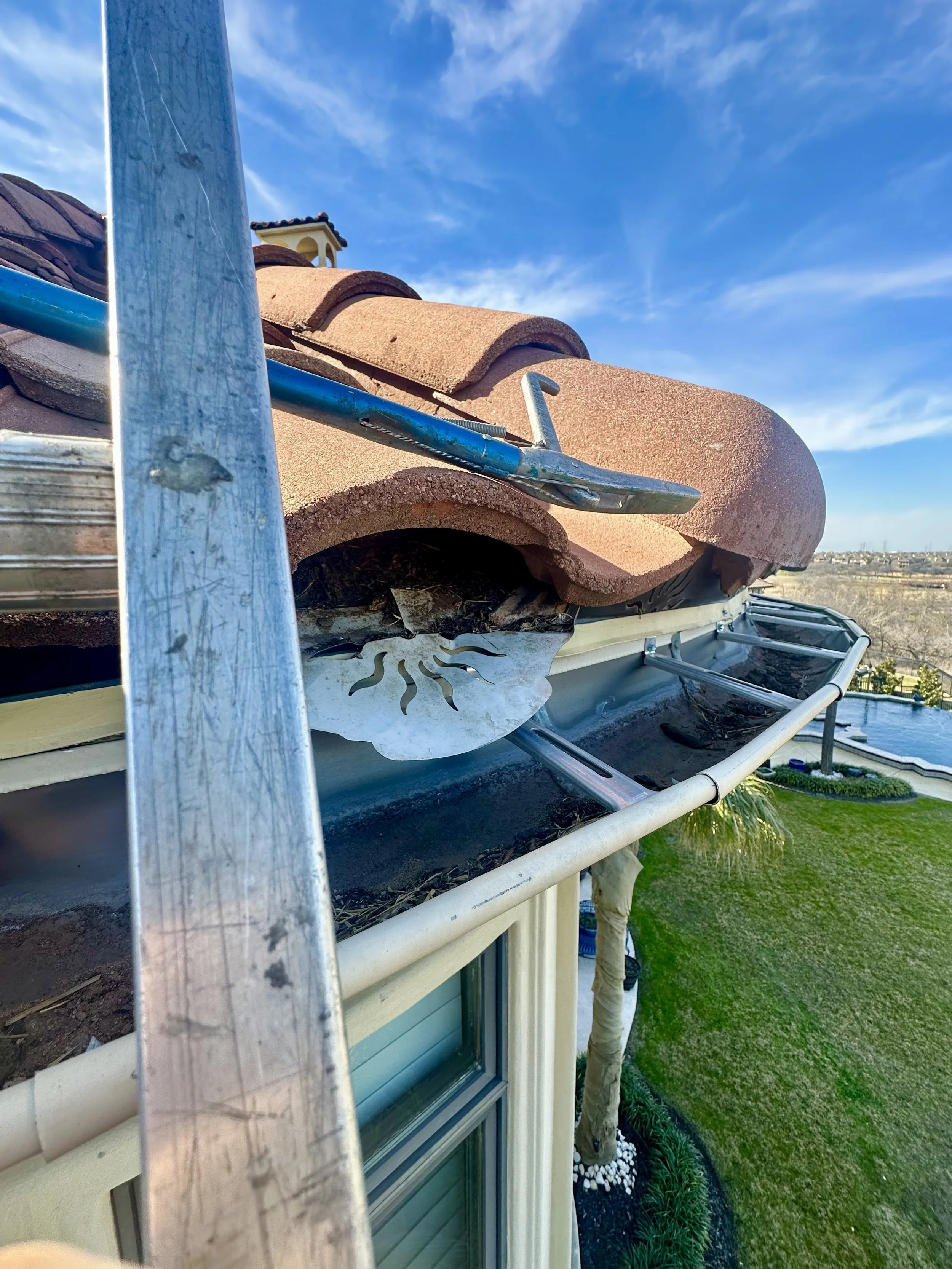 Roof with tiles, ladder, window, backyard with grass, trees, and pool under a blue sky with clouds.