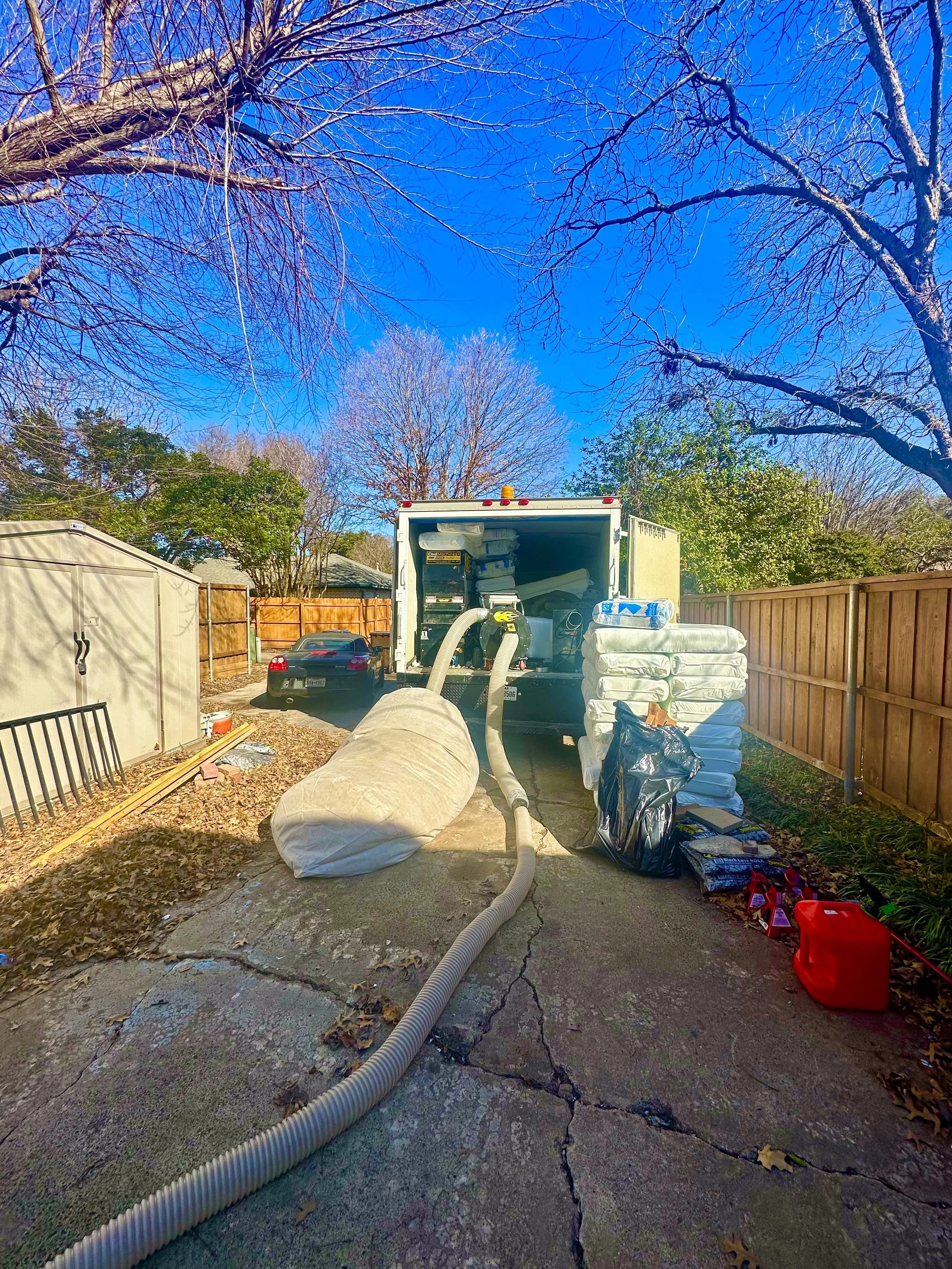 Backyard scene with a truck, ventilation hoses, stacks of insulation, tools, and a red gas can under a clear blue sky, trees without leaves, and a wooden fence. performing insulation removing and install