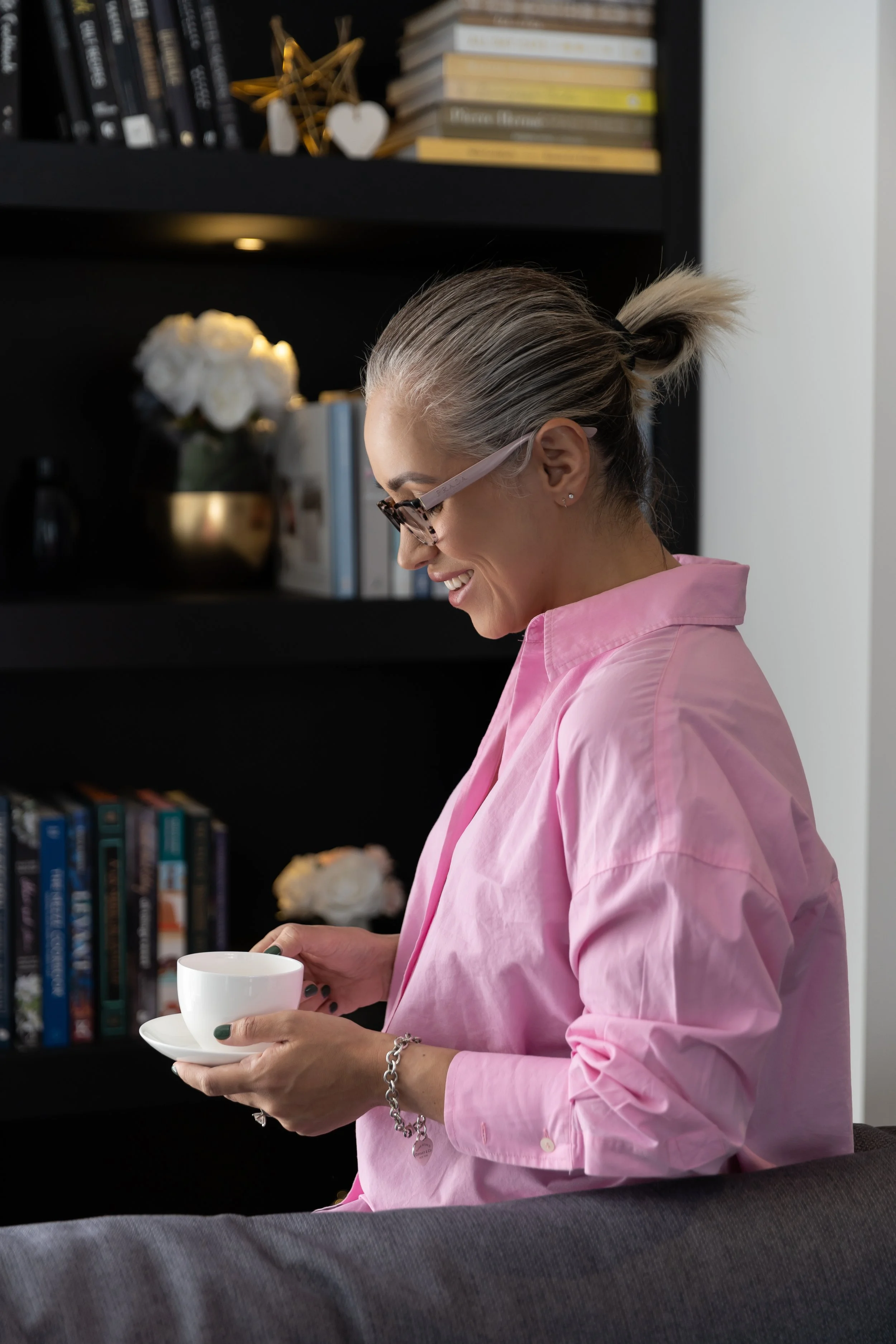 A woman with gray hair tied back, wearing glasses and a pink shirt, is smiling and holding a white teacup with a saucer. She is standing in a room with bookshelves and decorative items in the background.