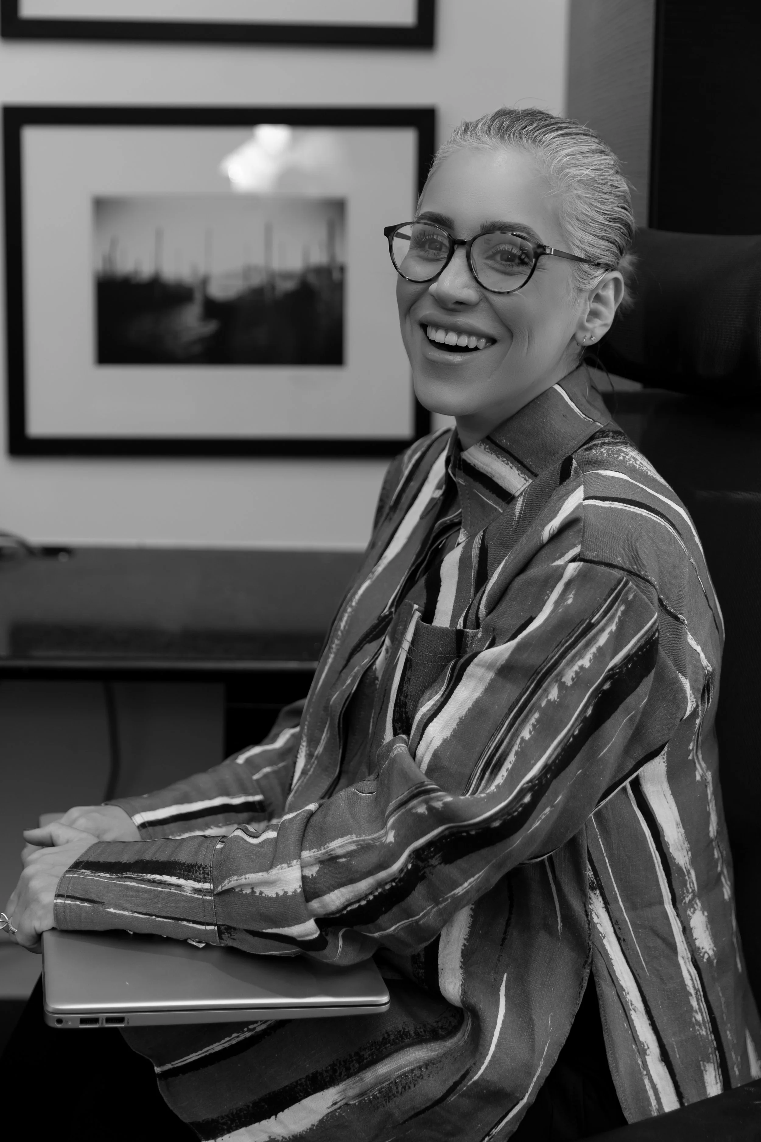 A woman with glasses and short, light-colored hair smiling and sitting at a table with a closed laptop in front of her. There are framed photographs on the wall behind her.
