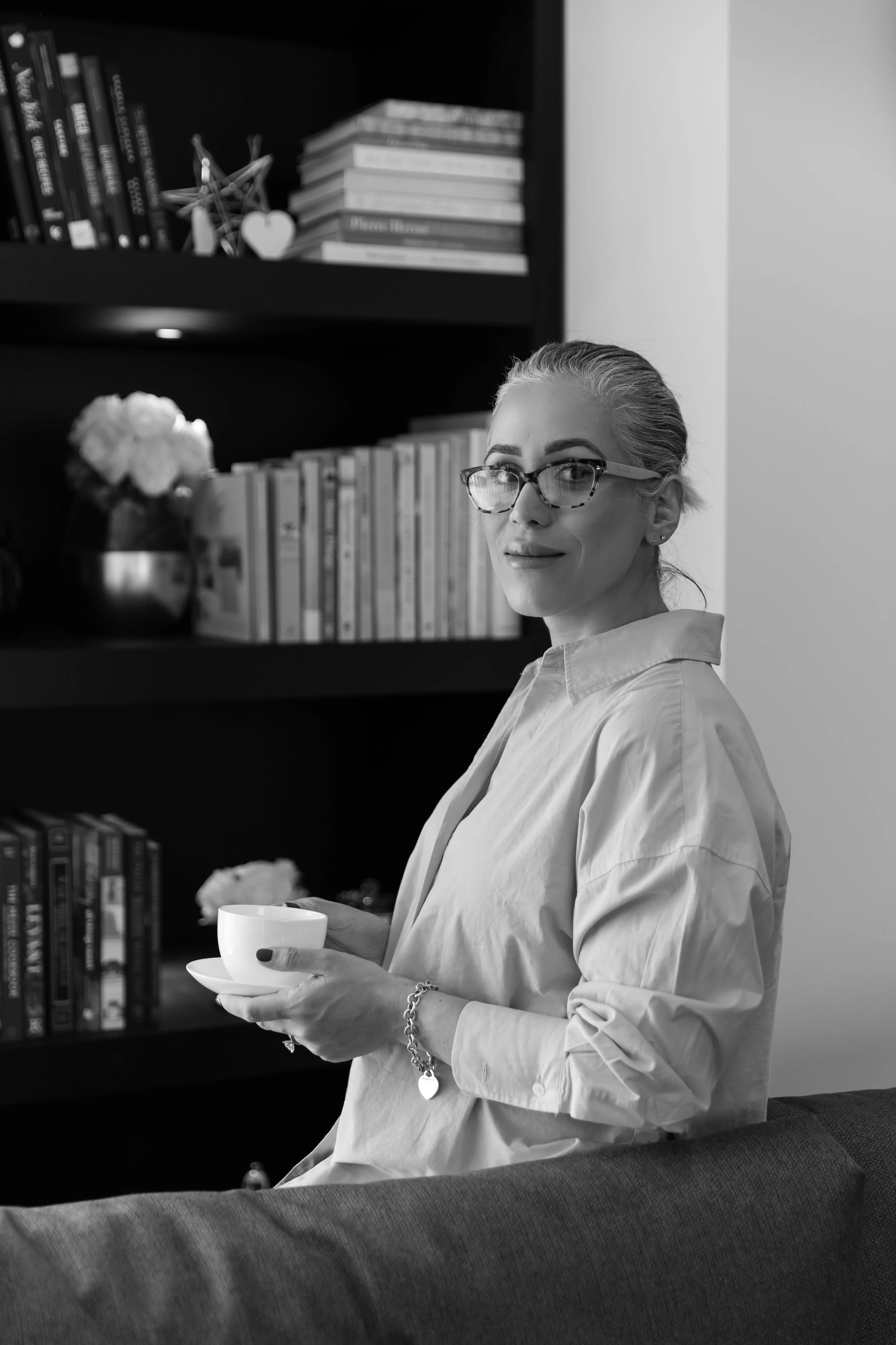 A woman with glasses holding a cup in a living room or library setting. She is standing in front of a bookshelf filled with books and decorative items. The photo is in black and white.