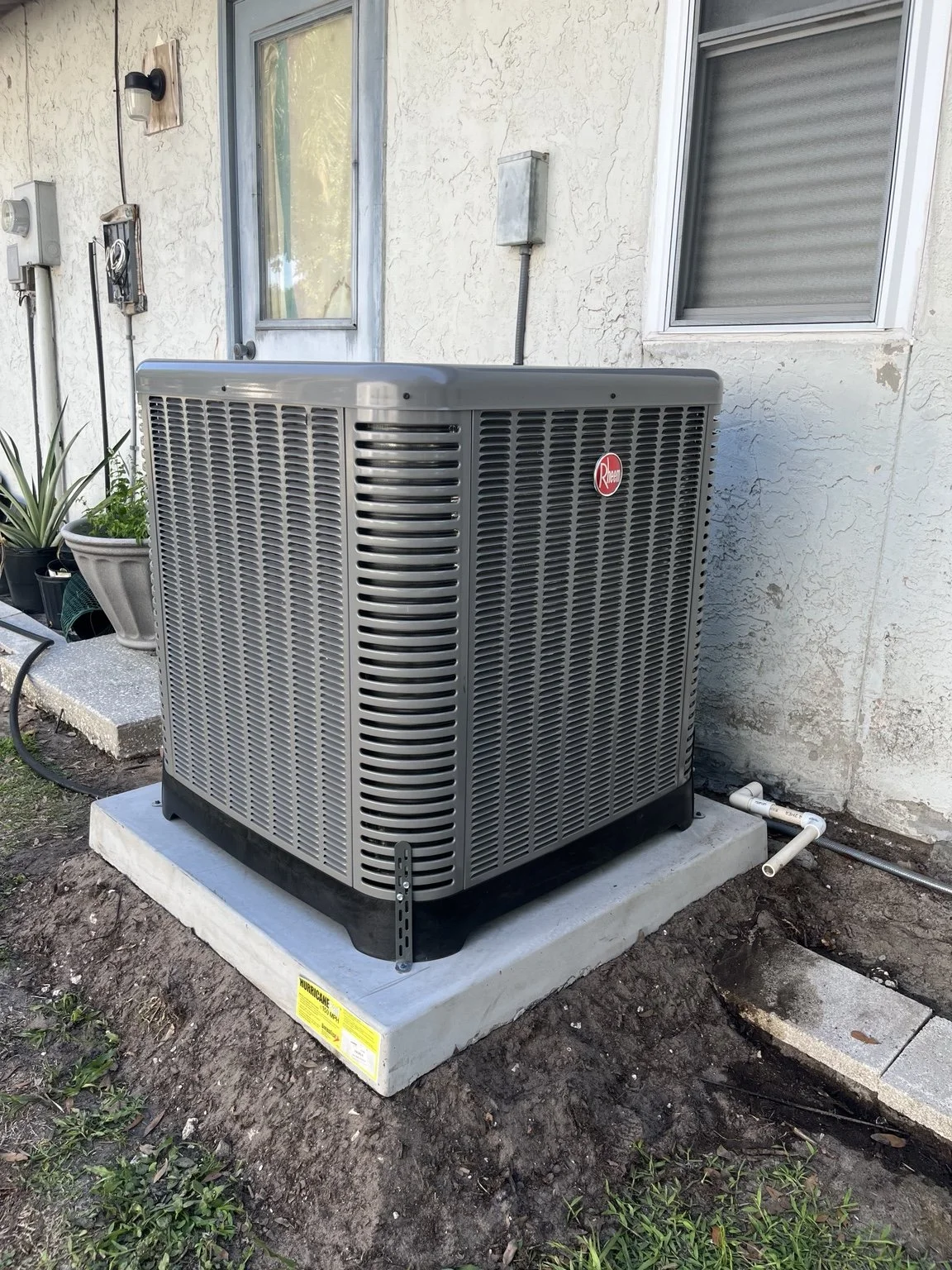 Outdoor temperature and air conditioning unit next to house wall, with plants nearby and exposed piping.