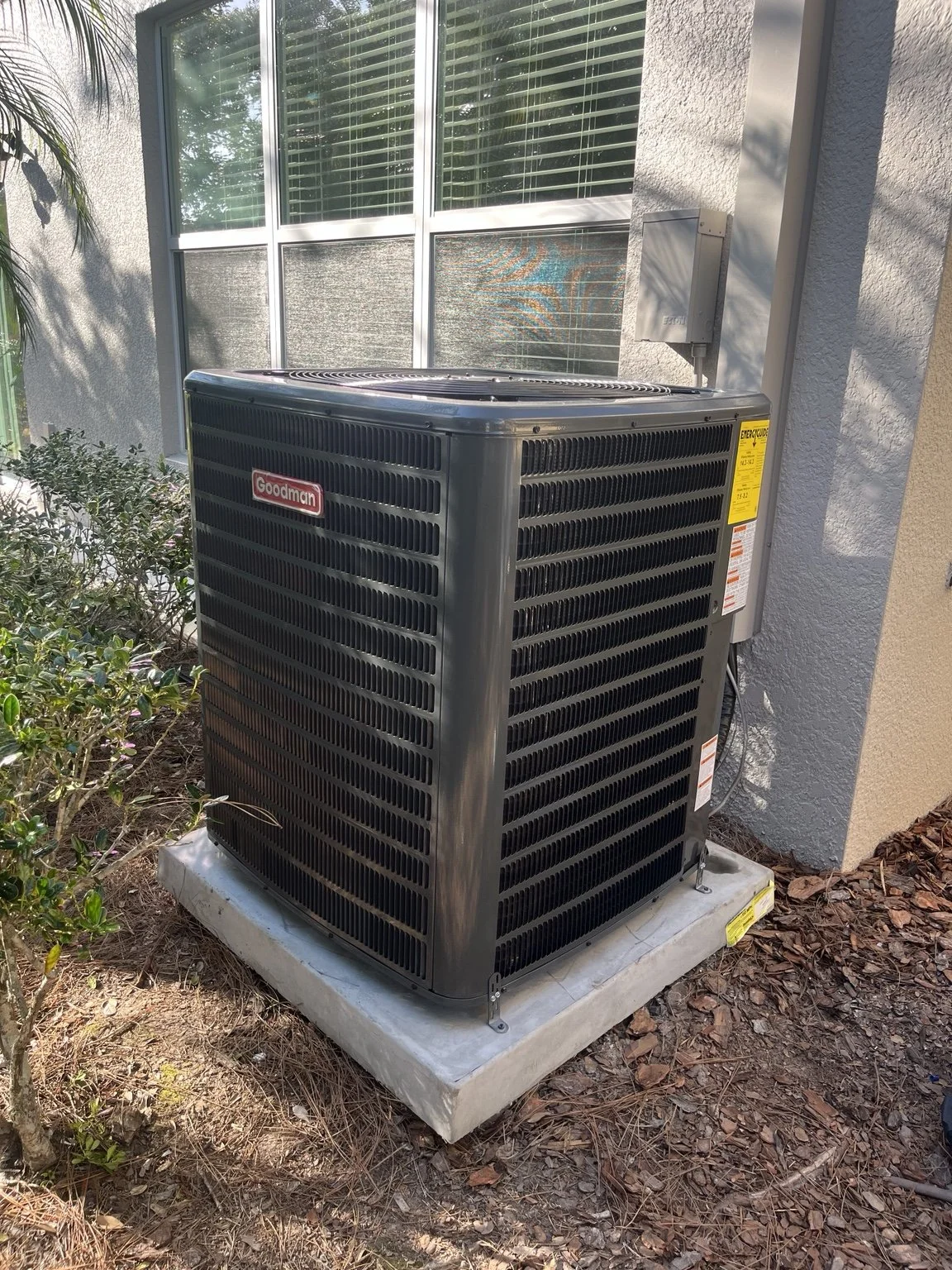 An outdoor Goodman air conditioning unit installed on a concrete pad next to a house wall and window, surrounded by mulch and small bushes.