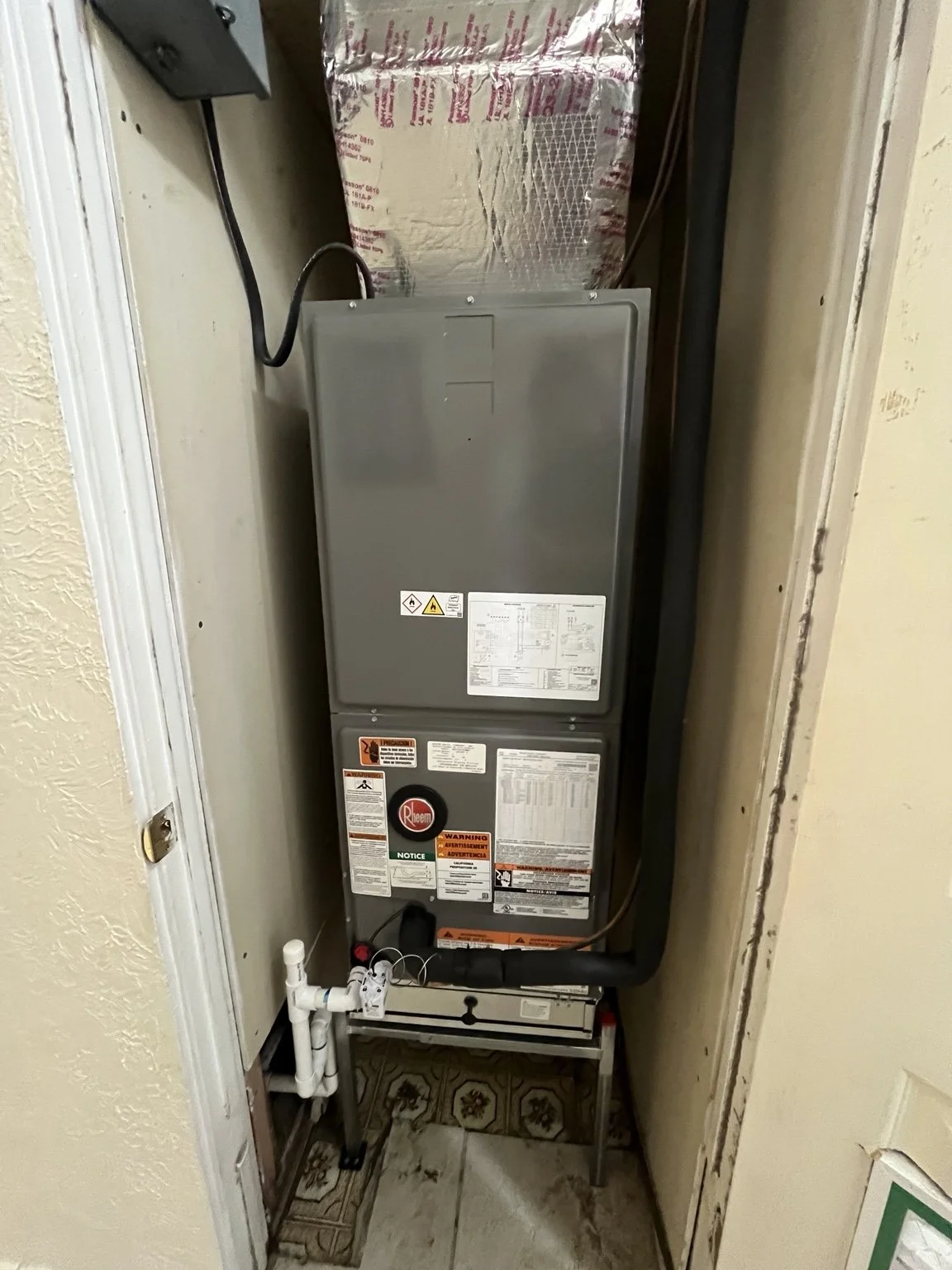 A utility closet containing a gray furnace or HVAC unit, with pipes and wiring, small space, beige walls, patterned tile flooring.